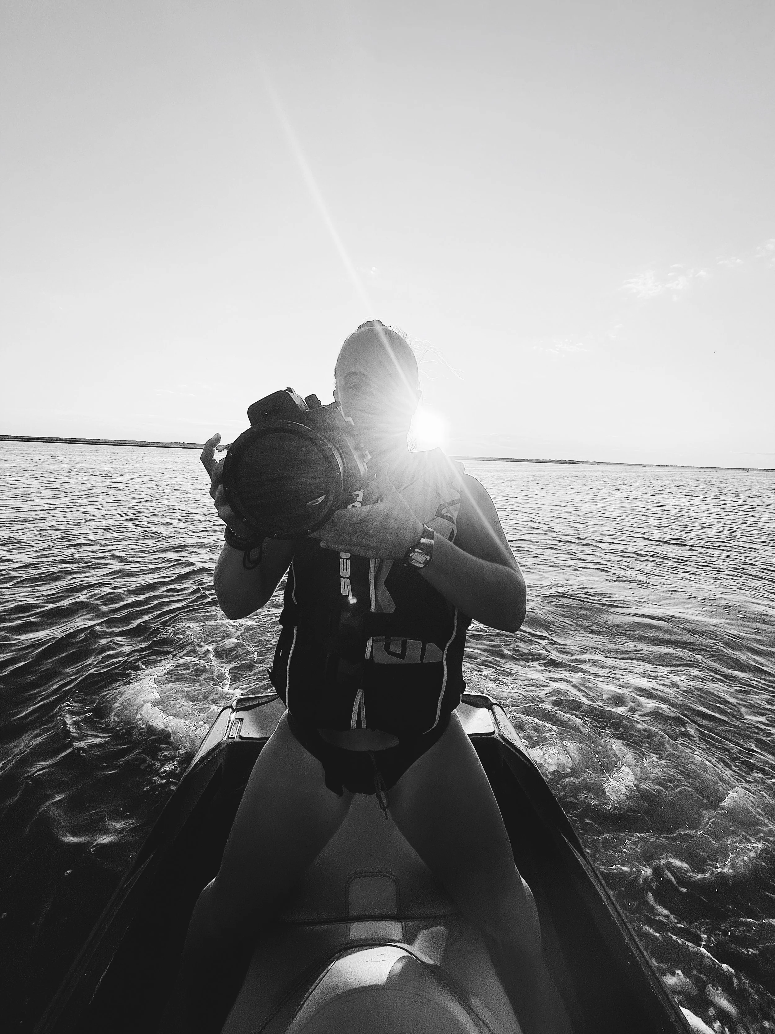 Person on a jet ski taking a photograph with a camera, facing the sunset over a body of water, with the sun behind their head.