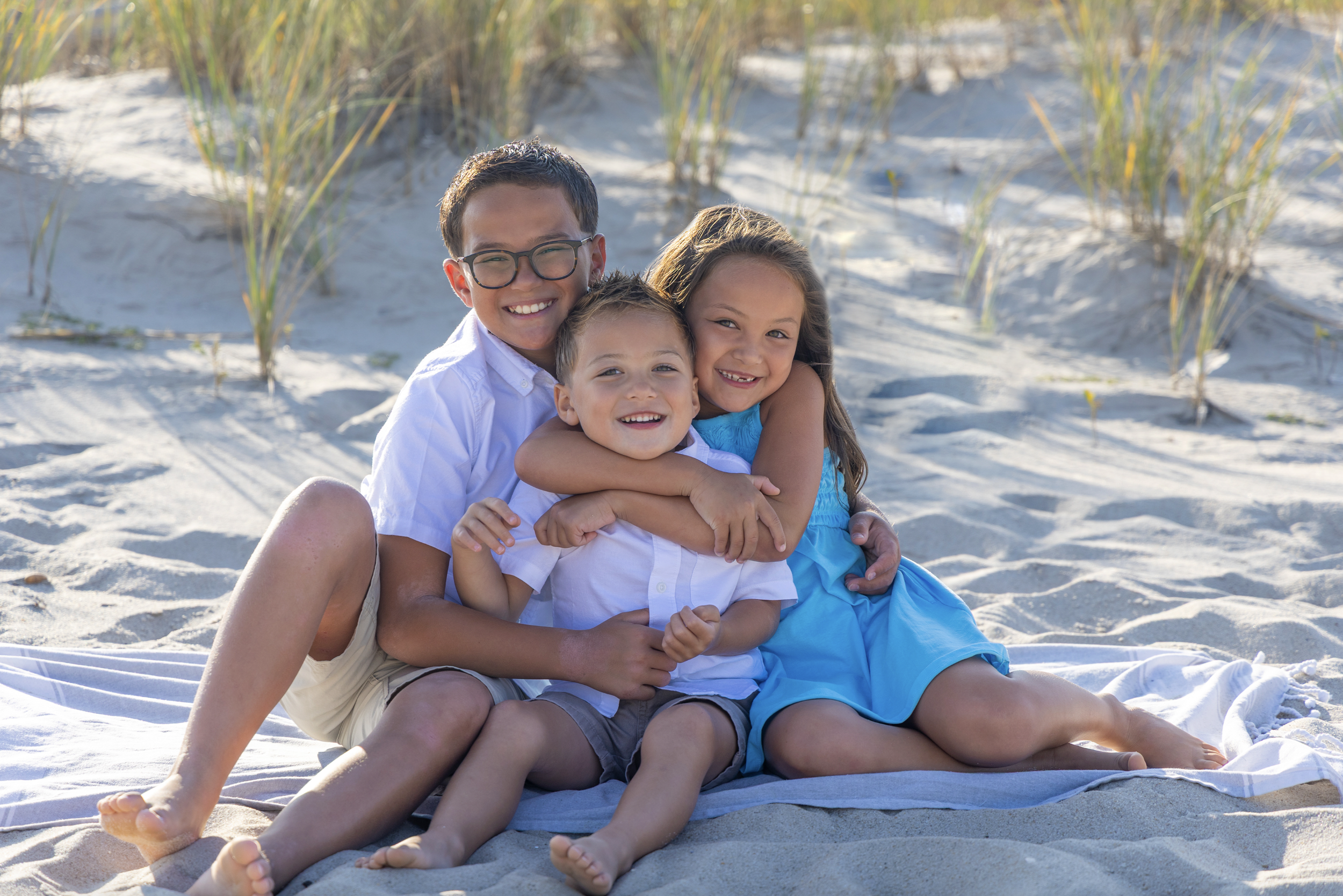 Four children smiling and hugging each other on a beach with sand and grasses in the background, wearing casual clothes.  Beach Family Photography Lavallette, New Jersey