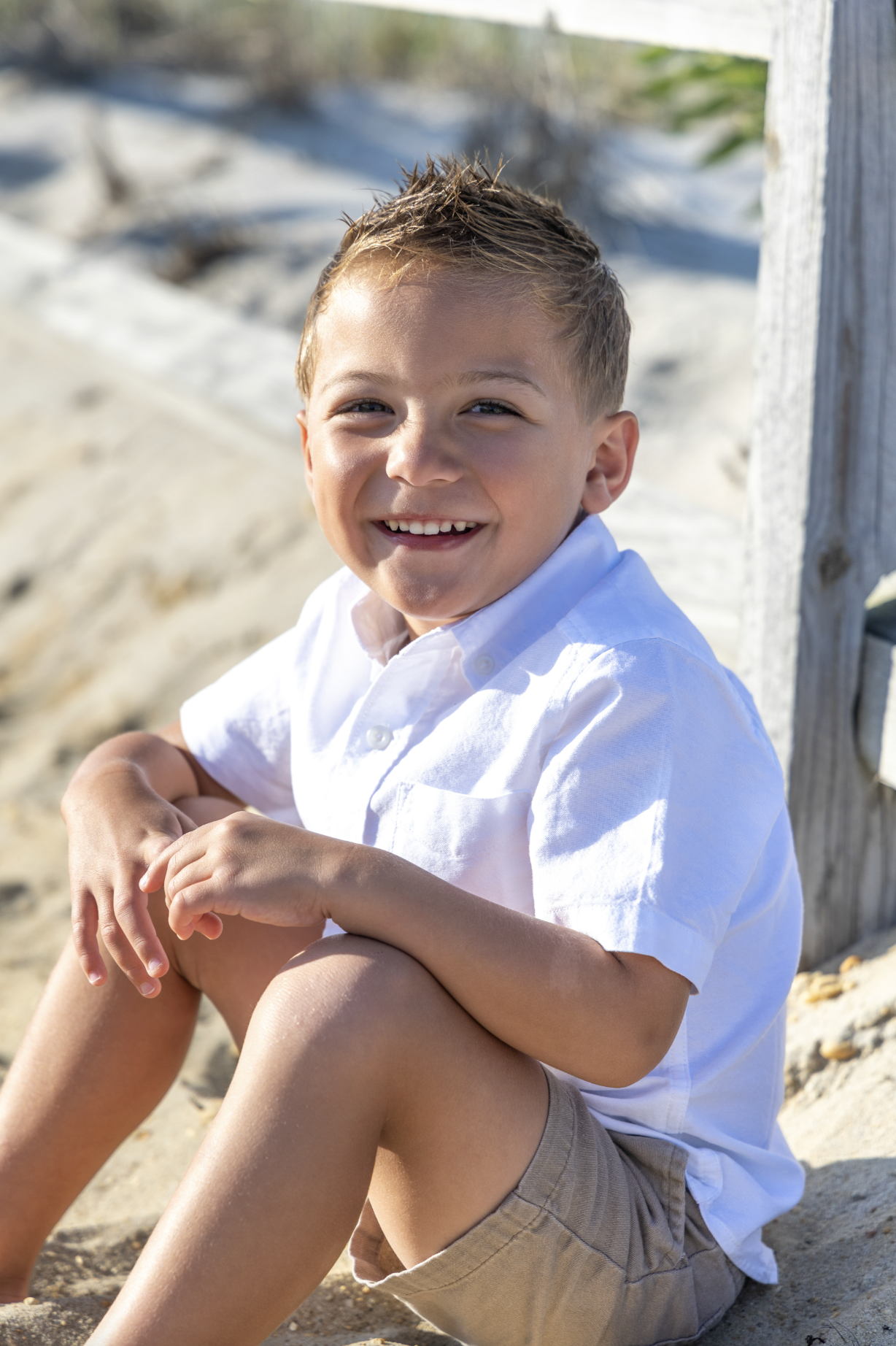 A young boy with short, spiky blond hair and a big smile, wearing a white shirt and khaki shorts, sitting on the sandy beach near a wooden post.