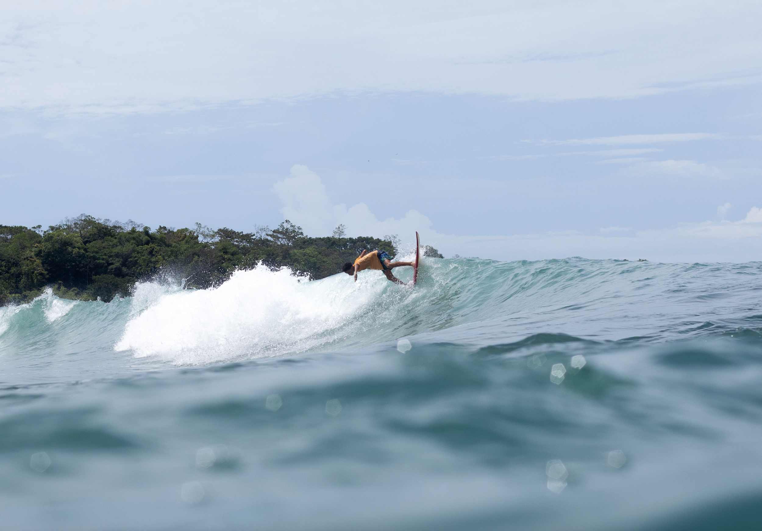 A person surfing on a blue wave near a tropical island with dense greenery.