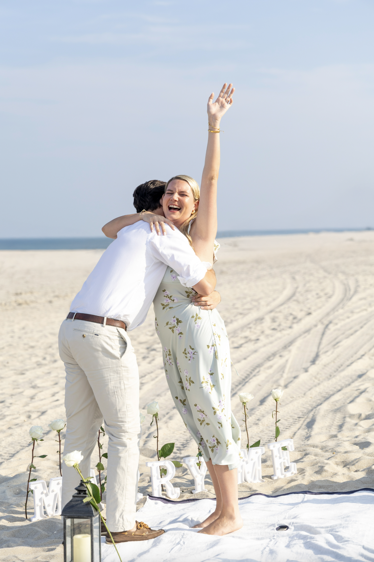 A couple on a beach wedding stands on a white cloth decorated with white roses and letters spelling 'MARRY ME.' The woman is smiling, raising her hand, and hugging the man, who is lightly kissing her on the cheek. The woman wears a white floral dress, and the man is in a white shirt and beige pants. The scene is cheerful and romantic on a sunny day with clear sky.