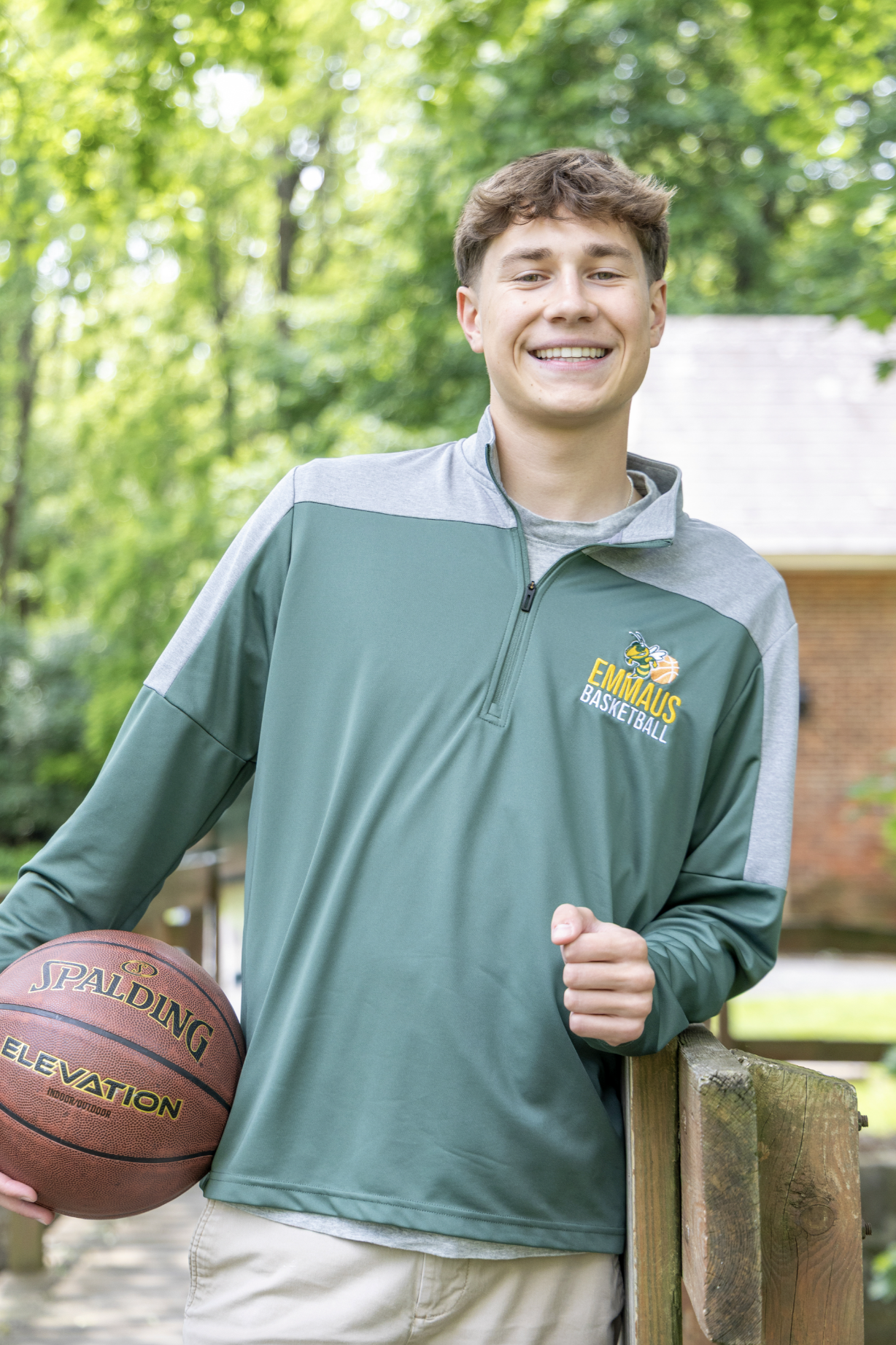 A young man smiling in a park, holding a basketball, wearing a green and gray 'Emmaus Basketball' jacket. High School Senior Graduation Photography