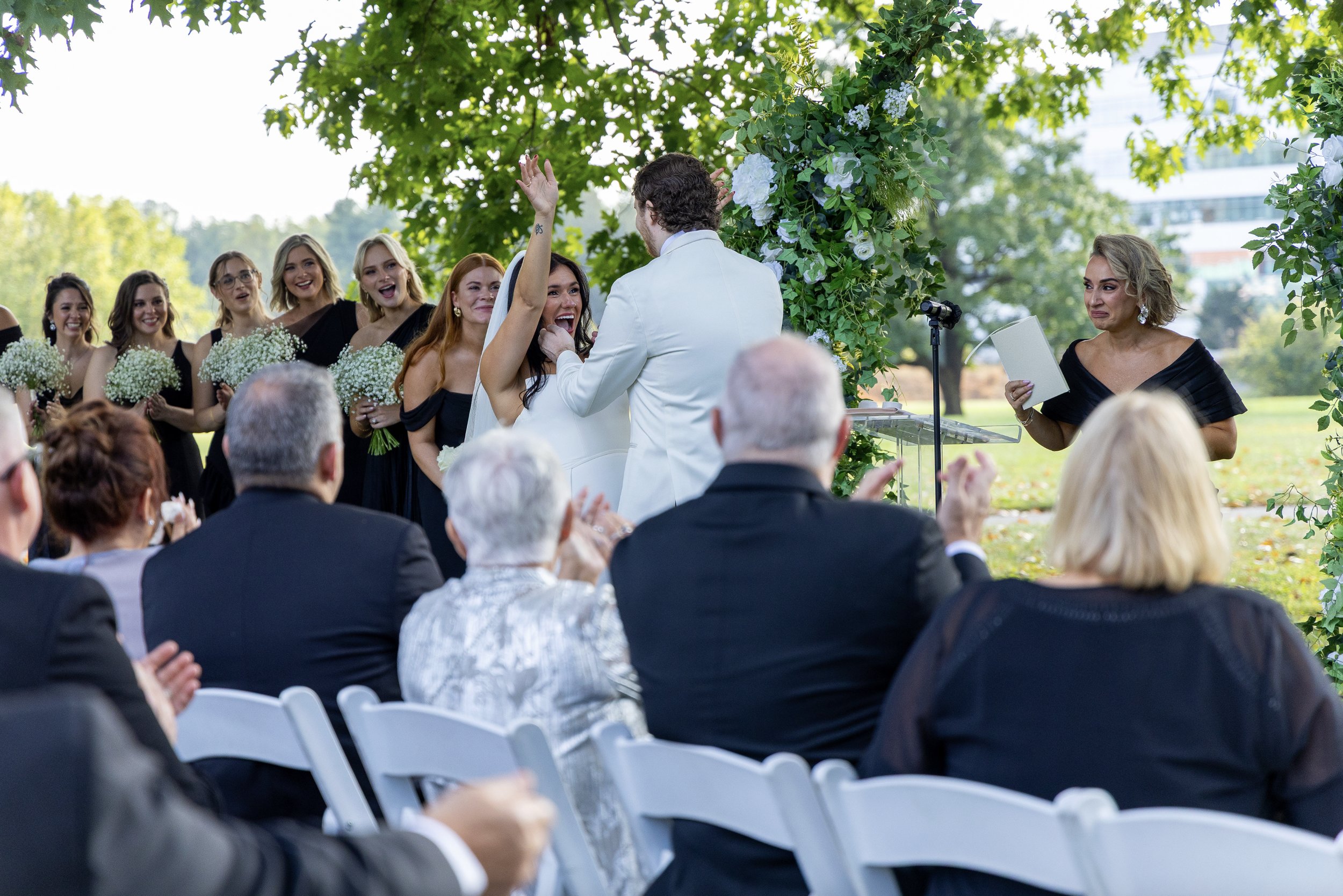 A wedding ceremony outdoors with the couple exchanging rings under a floral arch, surrounded by bridesmaids and guests.