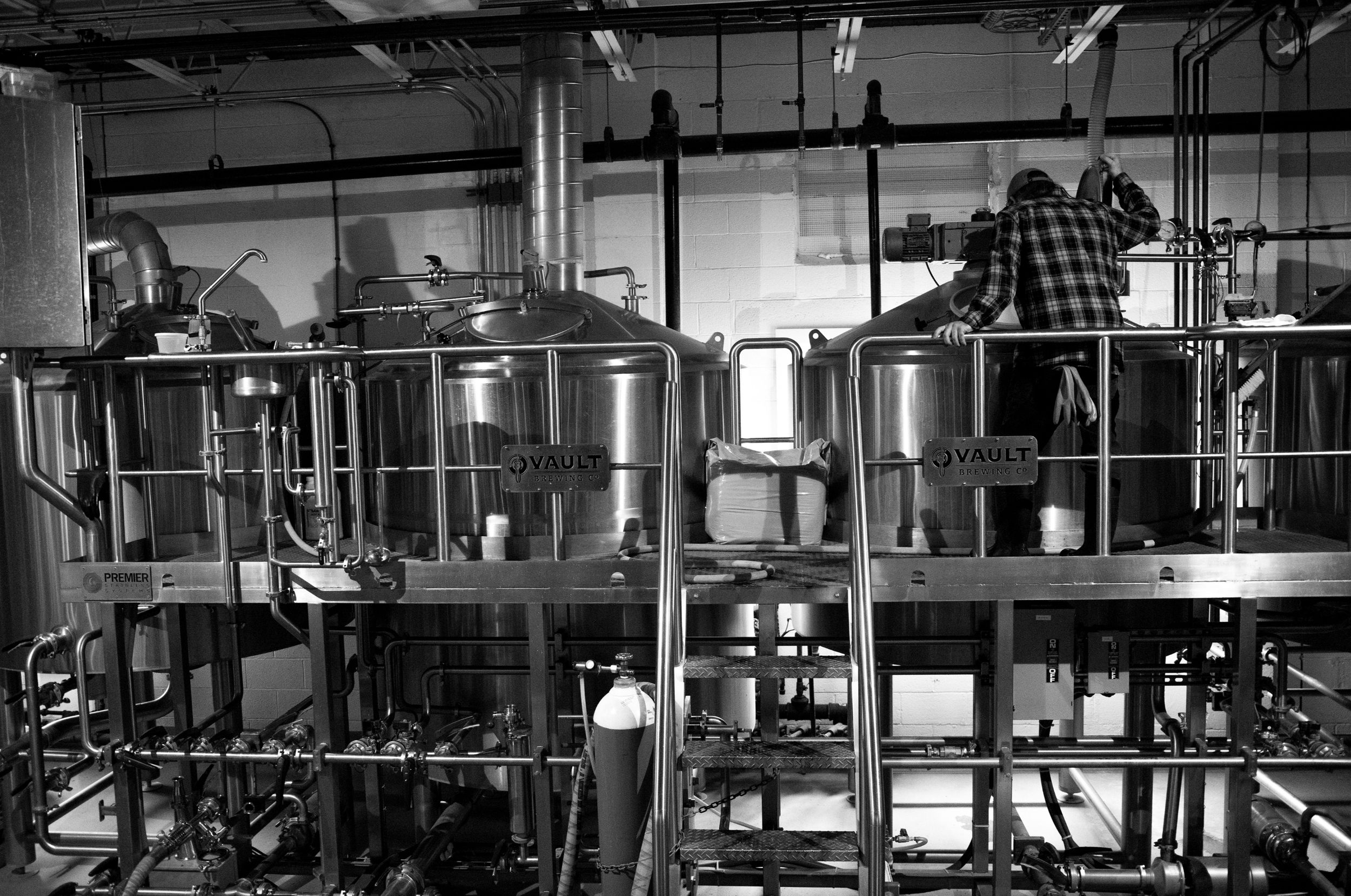 A man in a plaid shirt is working inside a brewery with large metal fermentation tanks, pipes, and industrial equipment. Vault Brewing, Yardley, Bucks County, Pennsylvania