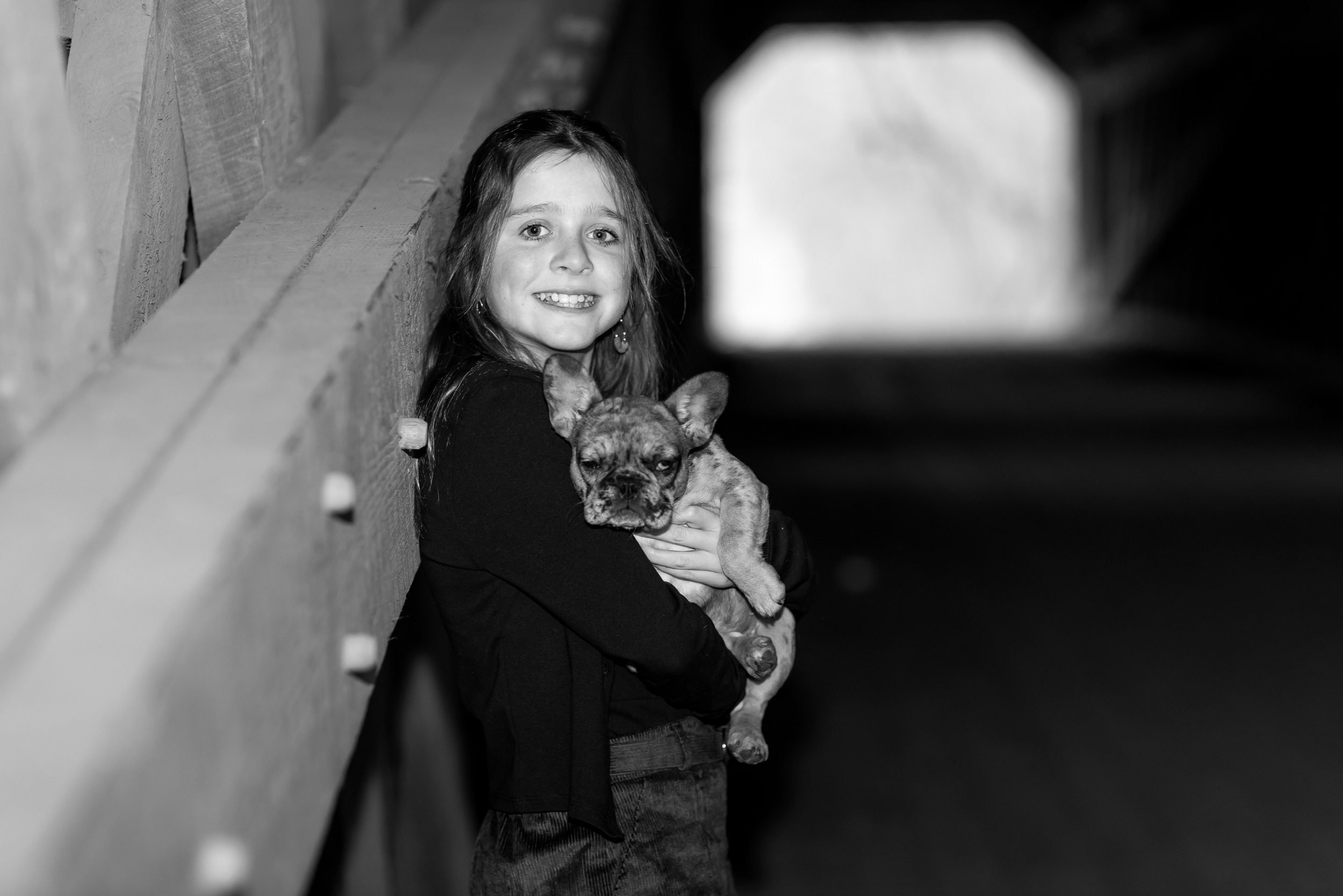 A young girl with long hair smiles while holding a small French Bulldog puppy in her arms indoors near a rustic wall, with a blurred background. Tyler State Park, Bucks County, New Jersey