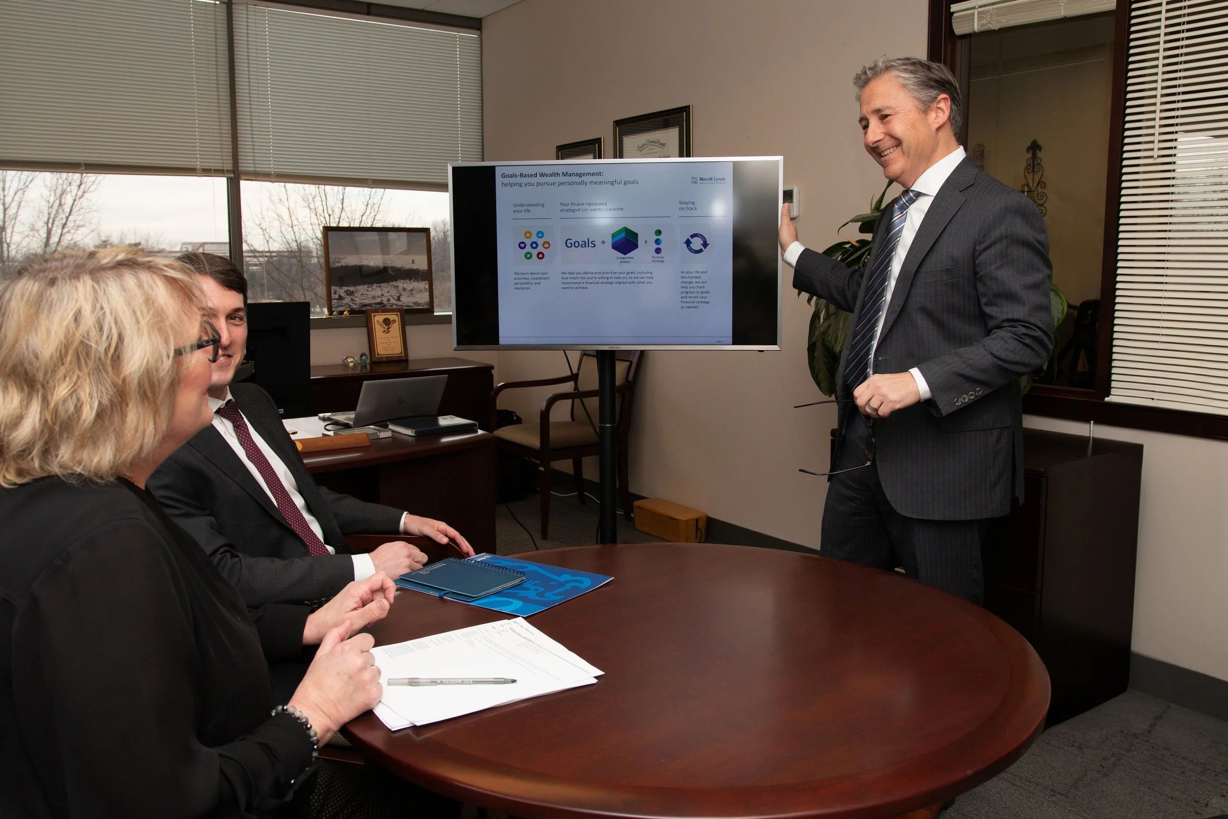 A businessman presenting a slide on goals to a group of three colleagues in a conference room, with a large screen displaying a presentation slide titled 'Goals-Based Wealth Management.' The room has large windows and some framed pictures on the wall.