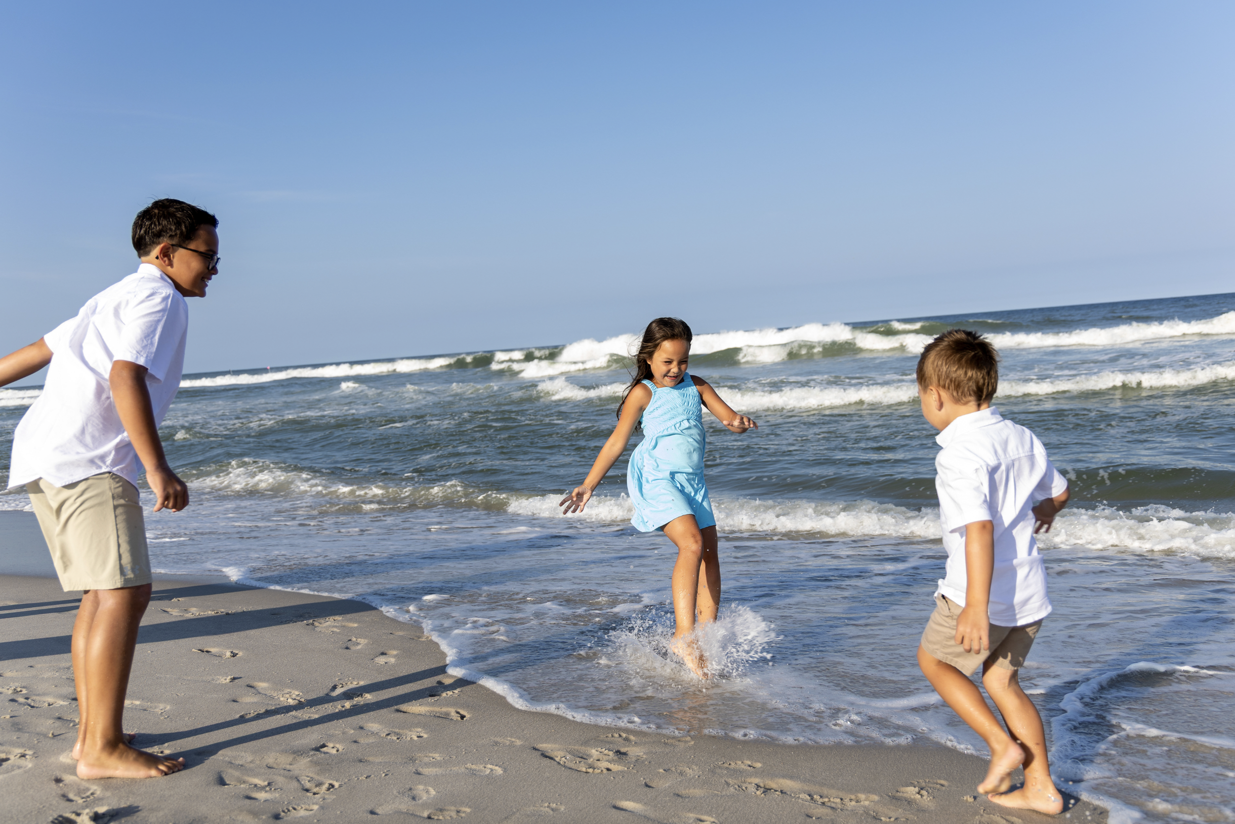 Four children playing and splashing in the ocean on a sandy beach under a clear blue sky.  Beach Family Photography Jersey Shore