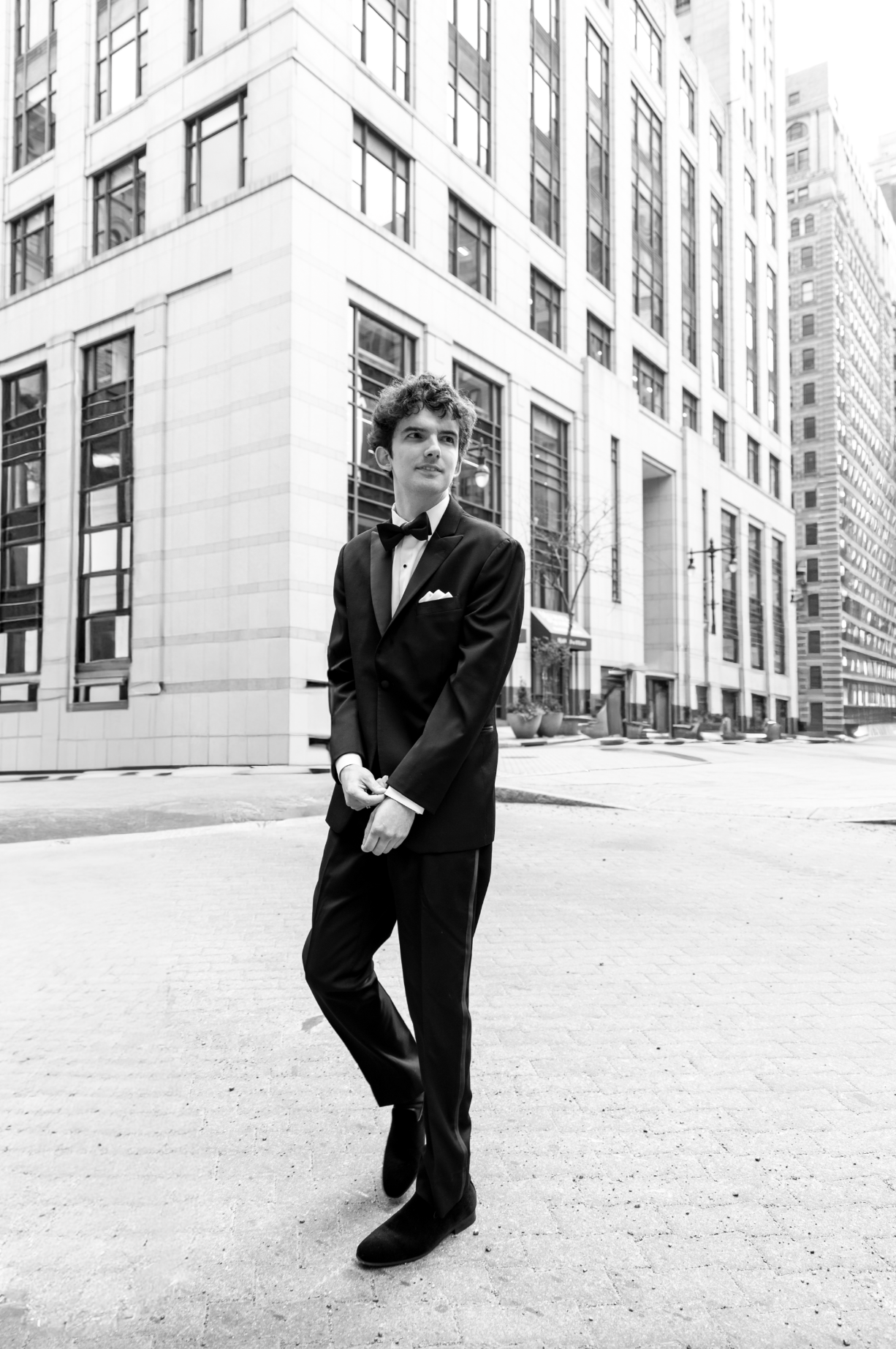 A young man in a tuxedo and bowtie walking on a city street in front of tall office buildings. philadelphia, PA, groom