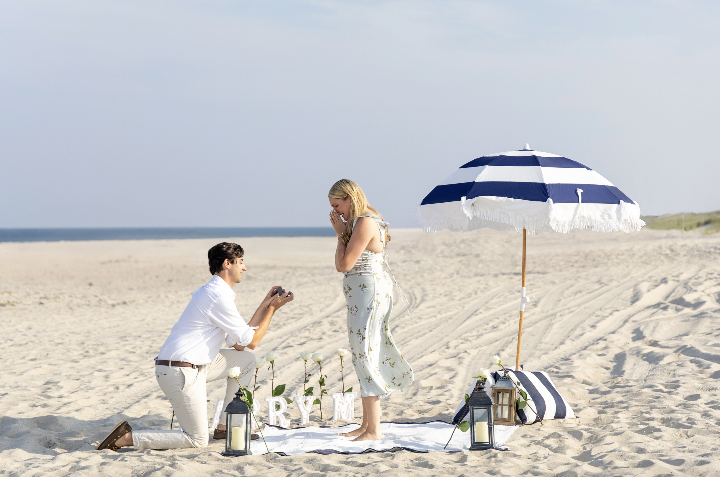 A man proposing marriage to a woman on a beach with a sailboat-themed setup, including a striped umbrella, lanterns, and flowers, during daytime. Lavallette, New Jersey Proposal