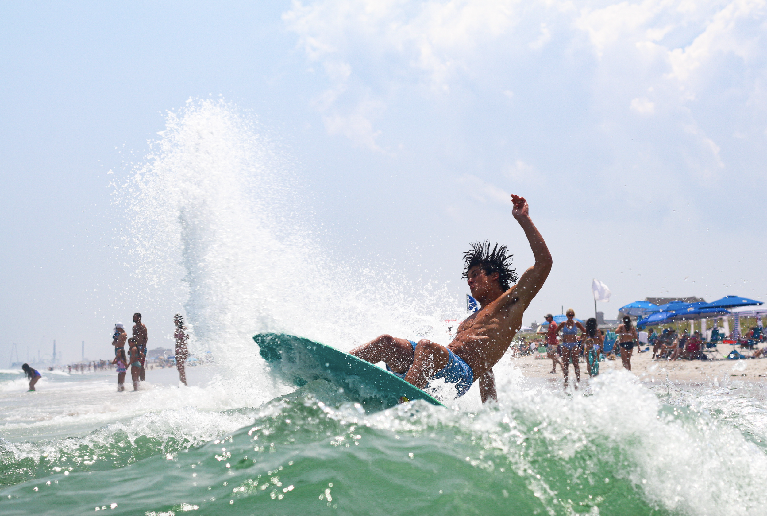A young man surfing in the ocean with a busy beach and blue umbrellas in the background.