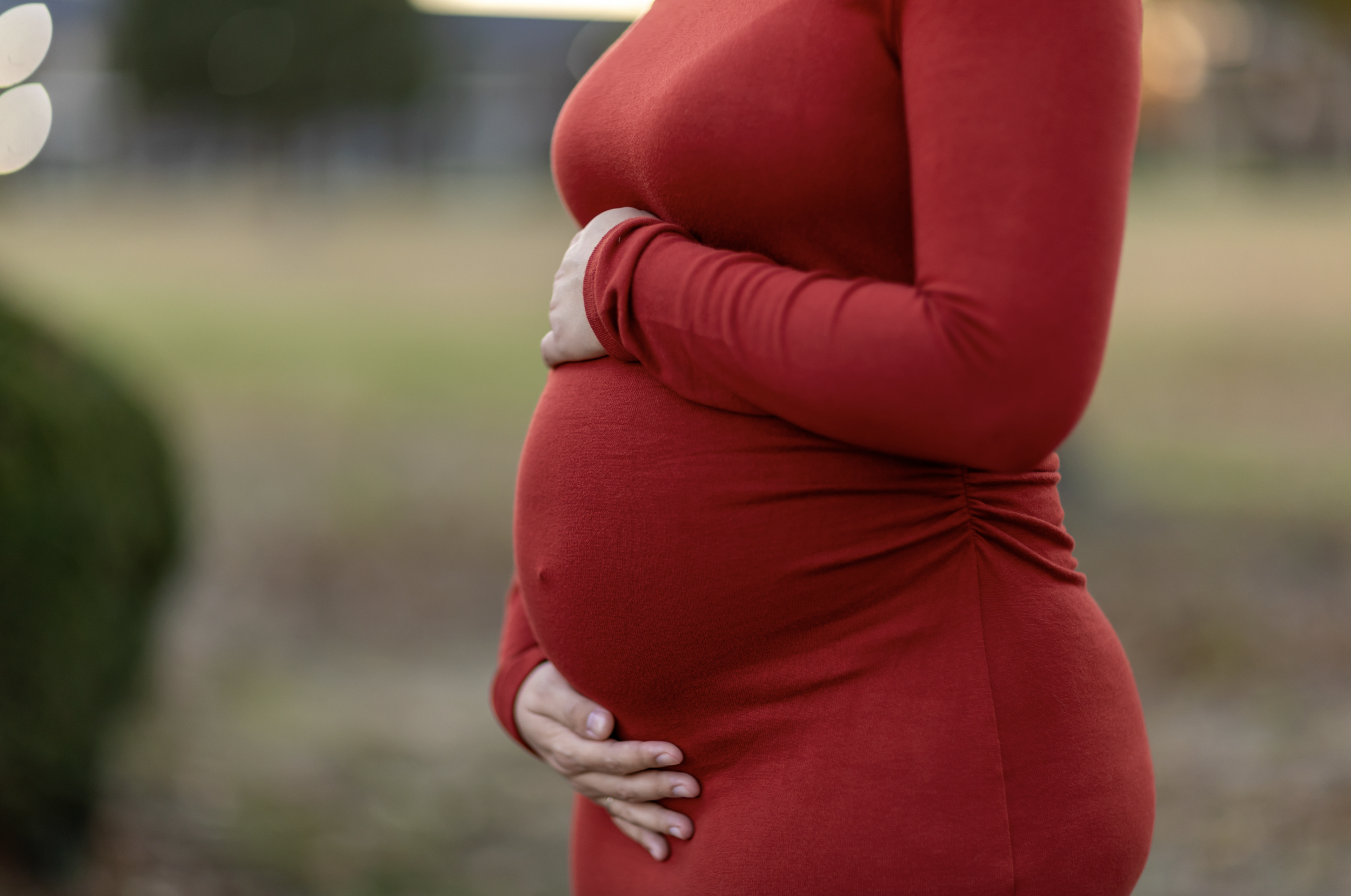 Close-up of a pregnant woman wearing a red long-sleeve dress, holding her belly with one hand, outdoors in blurred background. south new jersey maternity photography