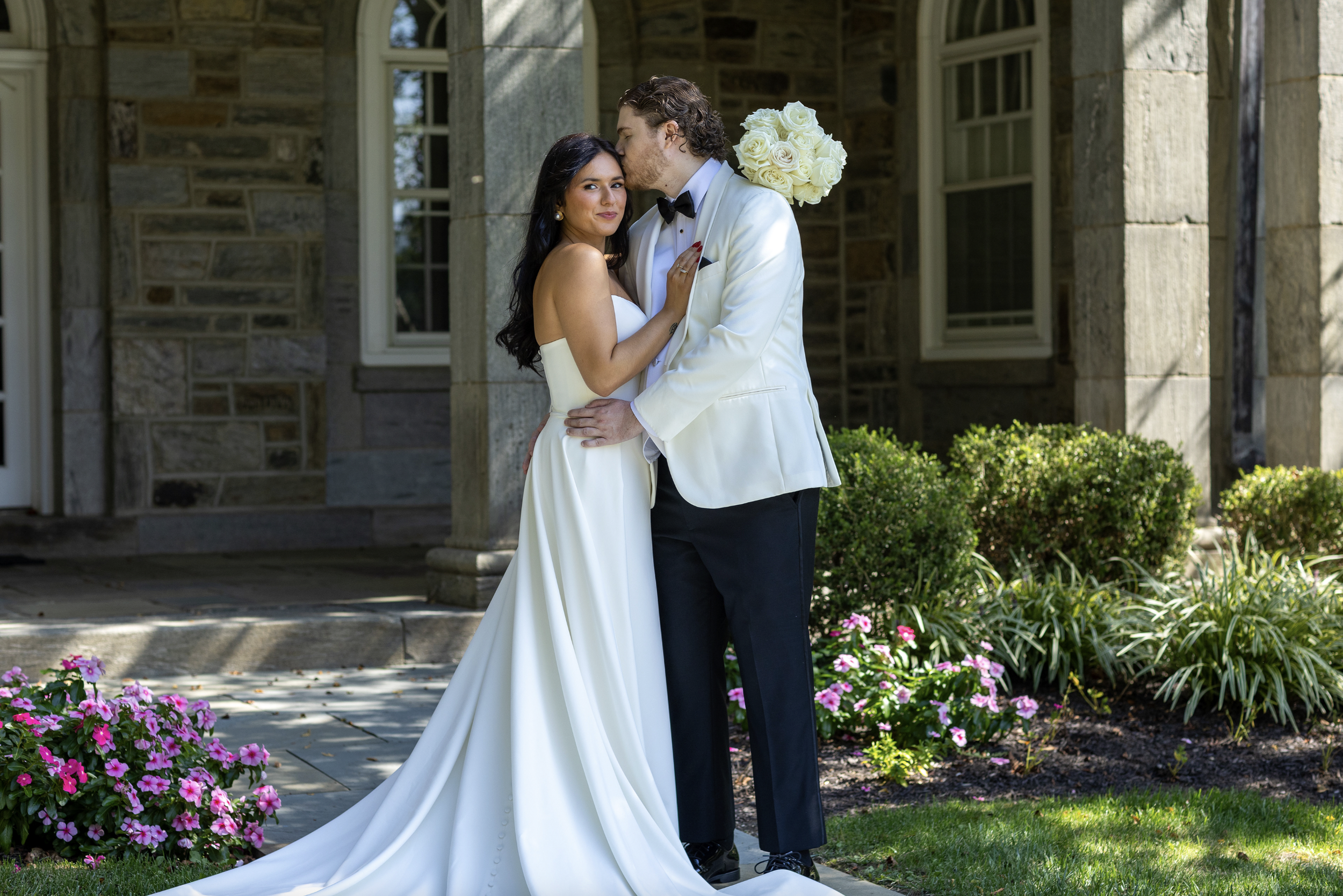 A newlywed couple stands outside a stone building, embracing and about to kiss, with flower beds and green shrubs in the background. New Jersey, Ocean County Wedding