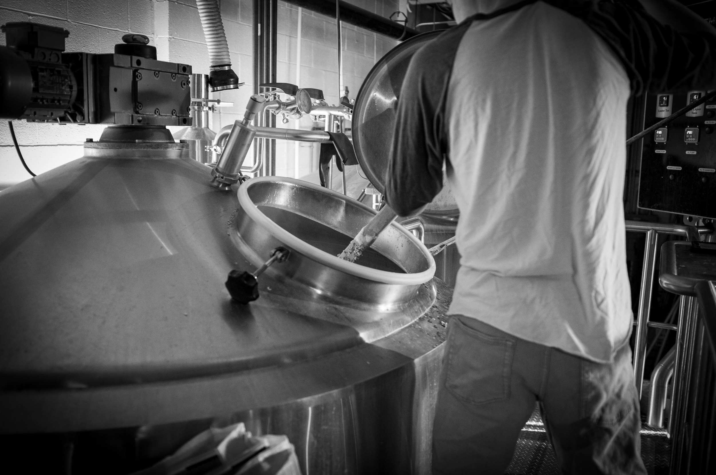 A person working with industrial equipment, possibly brewing or manufacturing, in a facility with stainless steel machinery and pipes, in black and white.