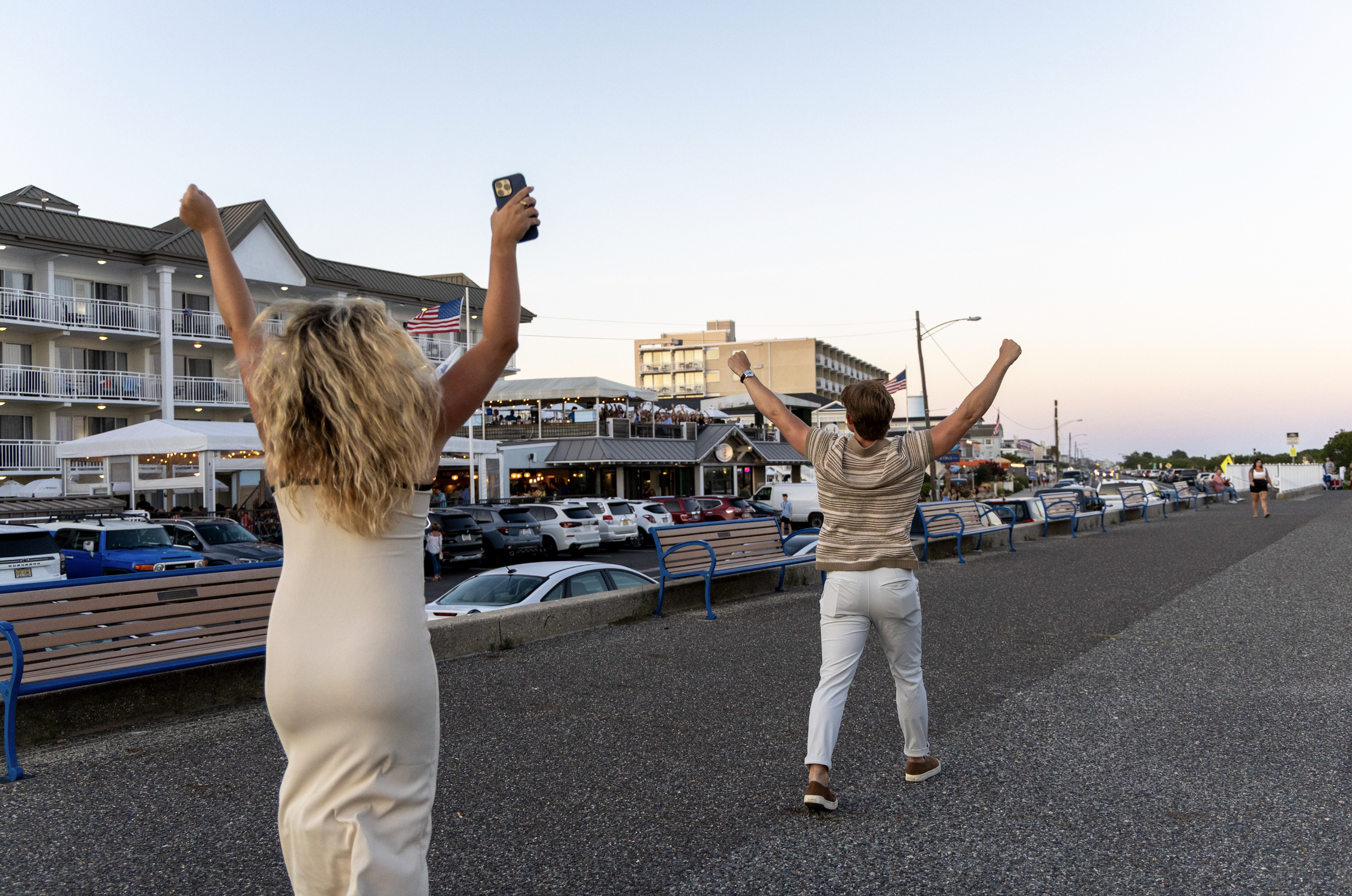 Two people celebrating by the seaside with arms raised, a woman taking a photo, dining and parking area, with buildings and American flags in the background at sunset. In Cape May, New Jersey.