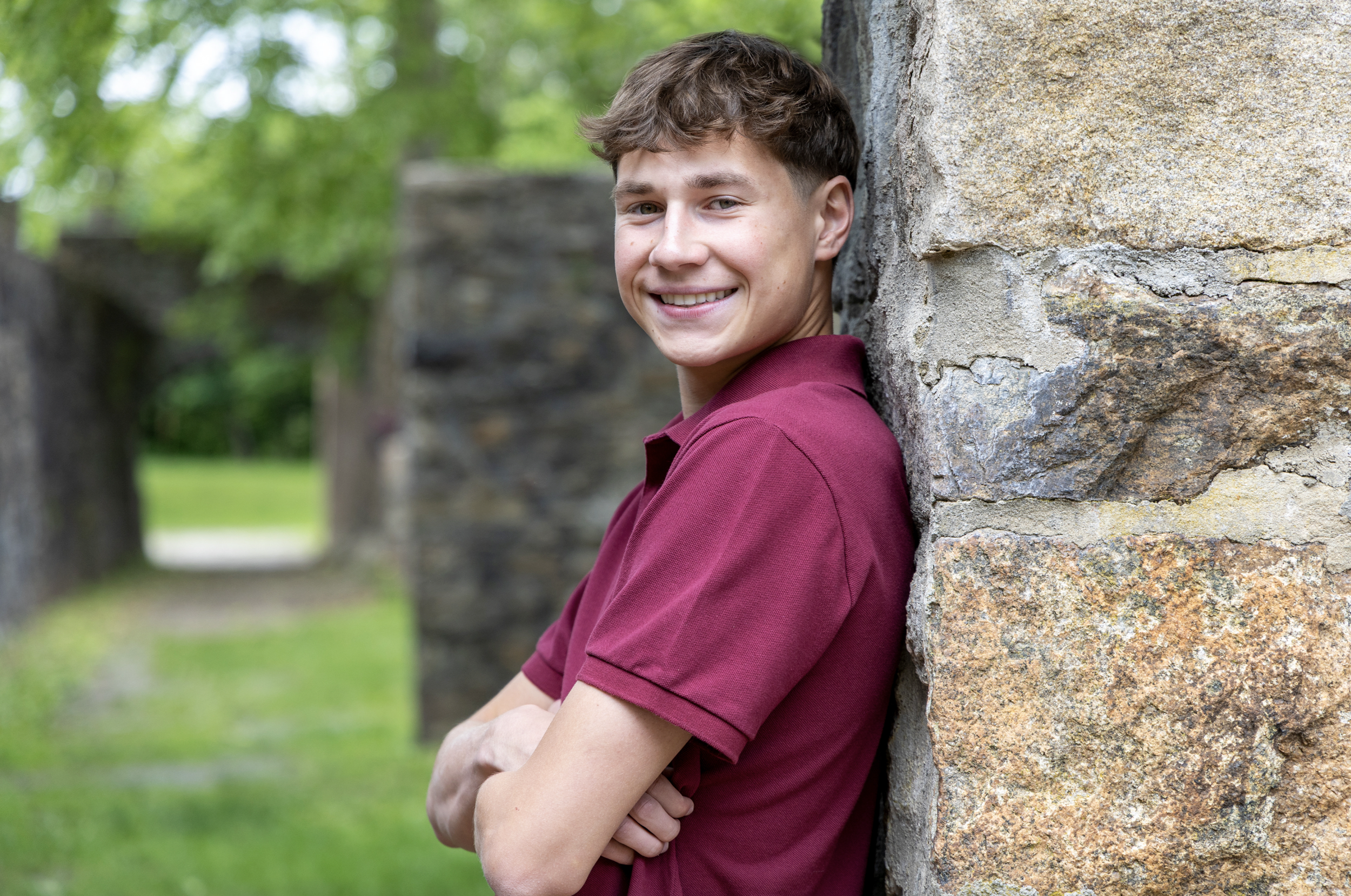 A young man with short brown hair smiling, wearing a maroon polo shirt, leaning against a stone wall outdoors surrounded by trees and greenery. Pennsylvania