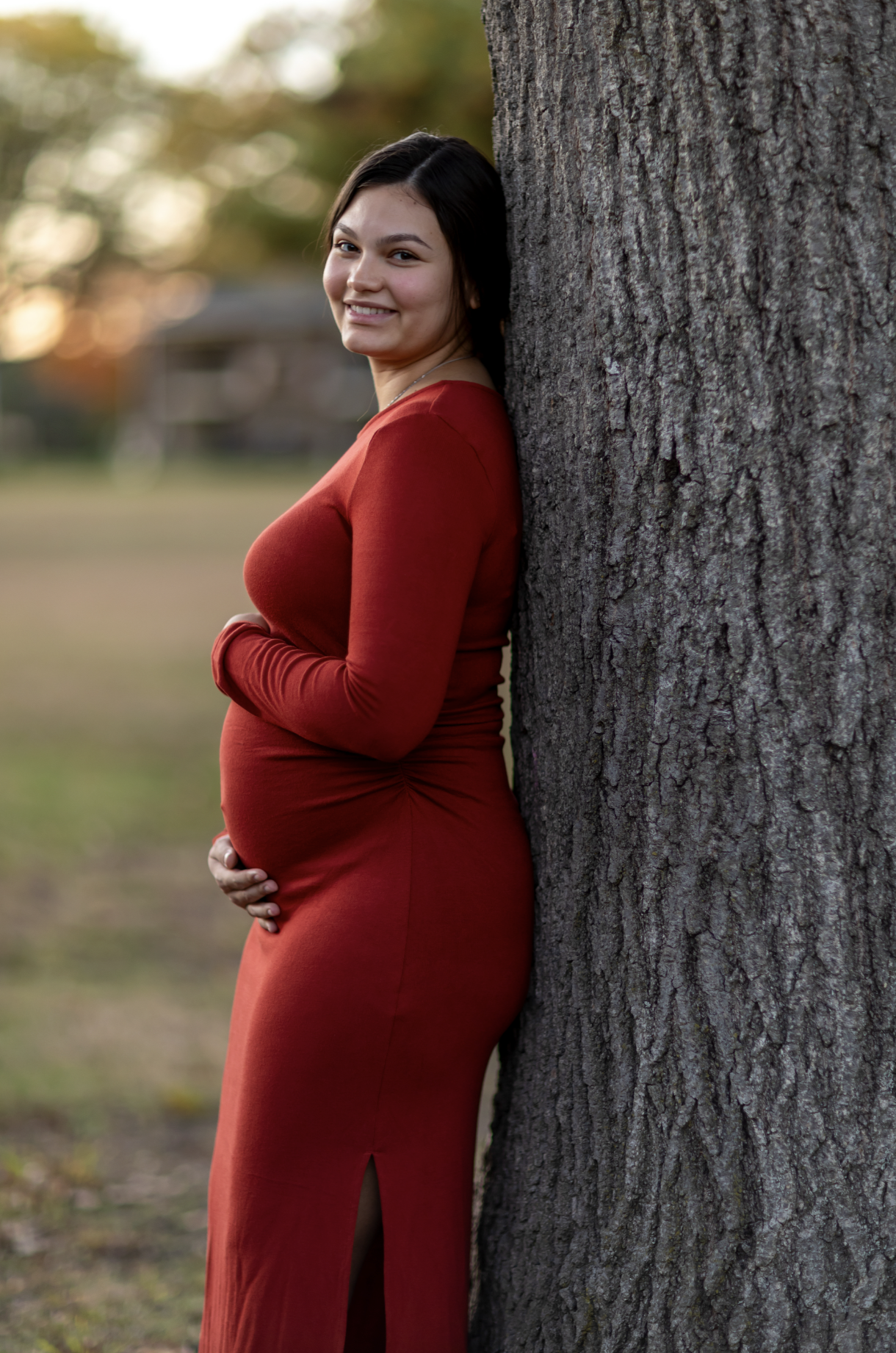 Pregnant woman in a red dress leaning against a tree outdoors, smiling at the camera during sunset.