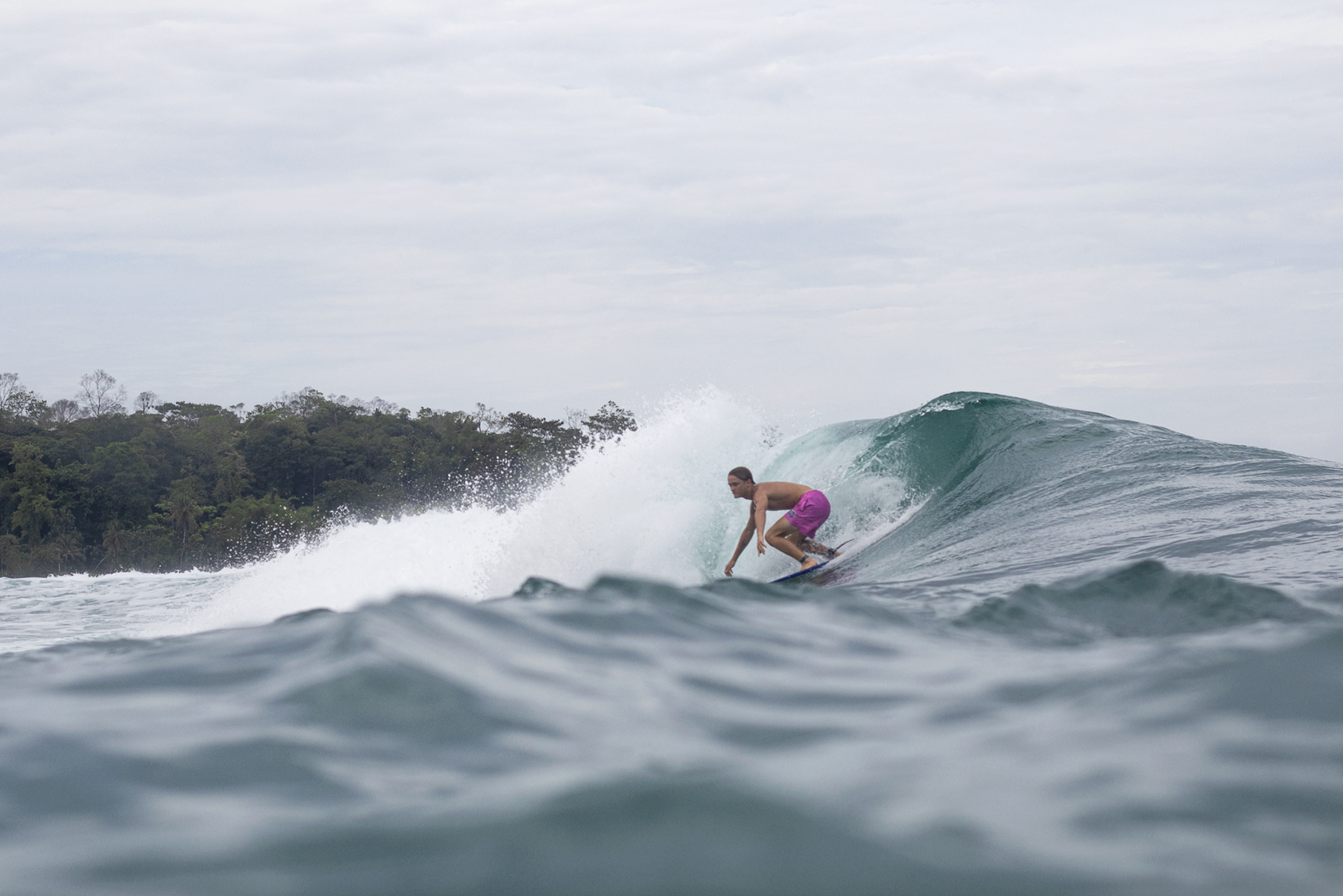 A person surfing on a wave in the ocean, wearing pink shorts with a backdrop of a cloudy sky and a forested shoreline.