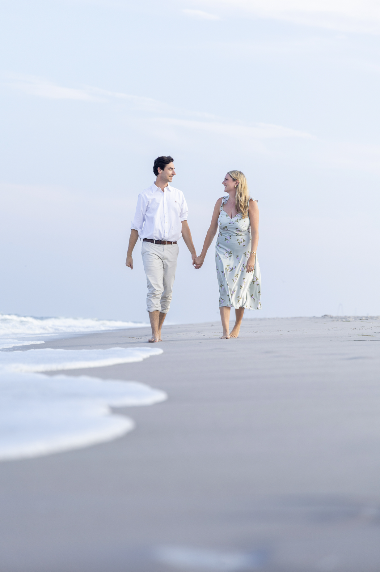 A couple walking hand-in-hand along the beach near the water, smiling at each other, with light clouds in the sky for an engagement session.