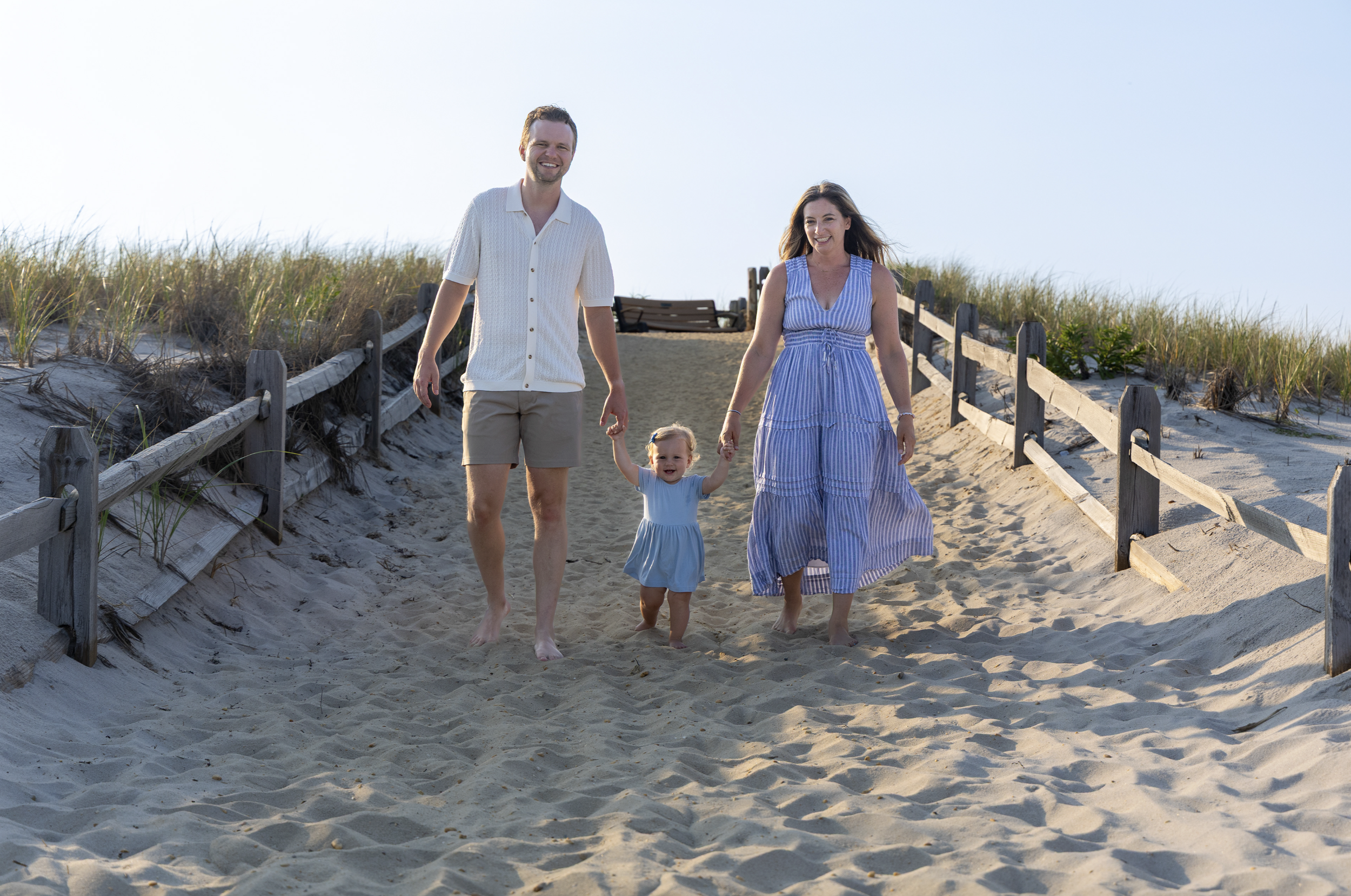 A family of three walking barefoot on a sandy beach path, holding hands, with dunes and grass in the background, enjoying a sunny day. Ocean County, New Jersey beach family photographer