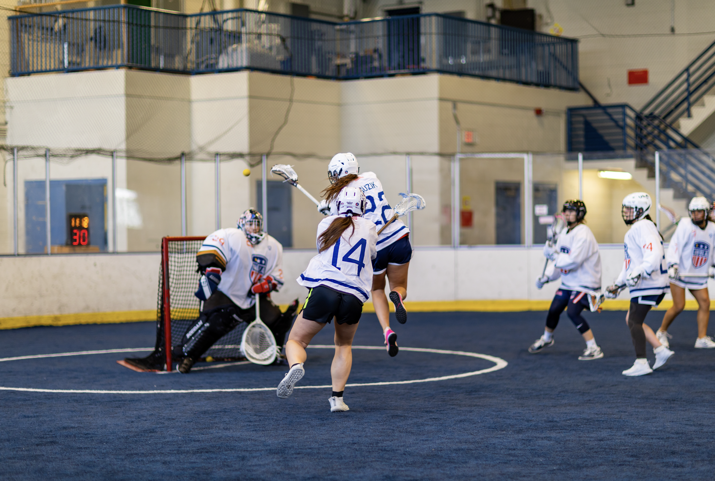 Women playing indoor lacrosse with a goalie defending the goal.