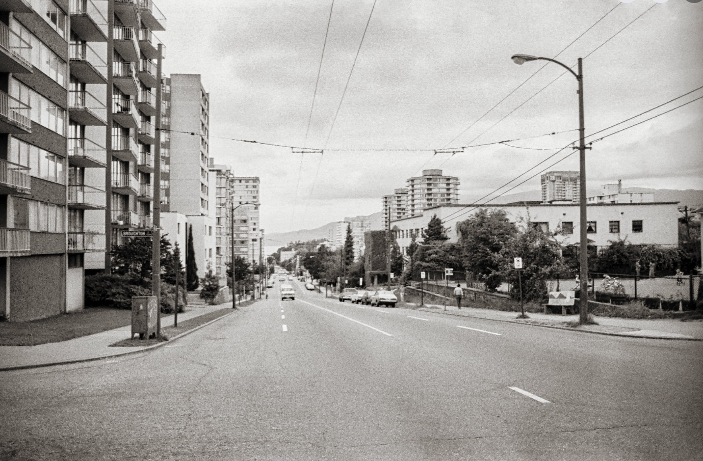 A black and white photo of an urban street with tall apartment buildings on the left side and shorter buildings on the right. Cars are parked along the street, and a few pedestrians walk on the sidewalks. Overhead tram wires stretch across the street, with a streetlamp on the right.