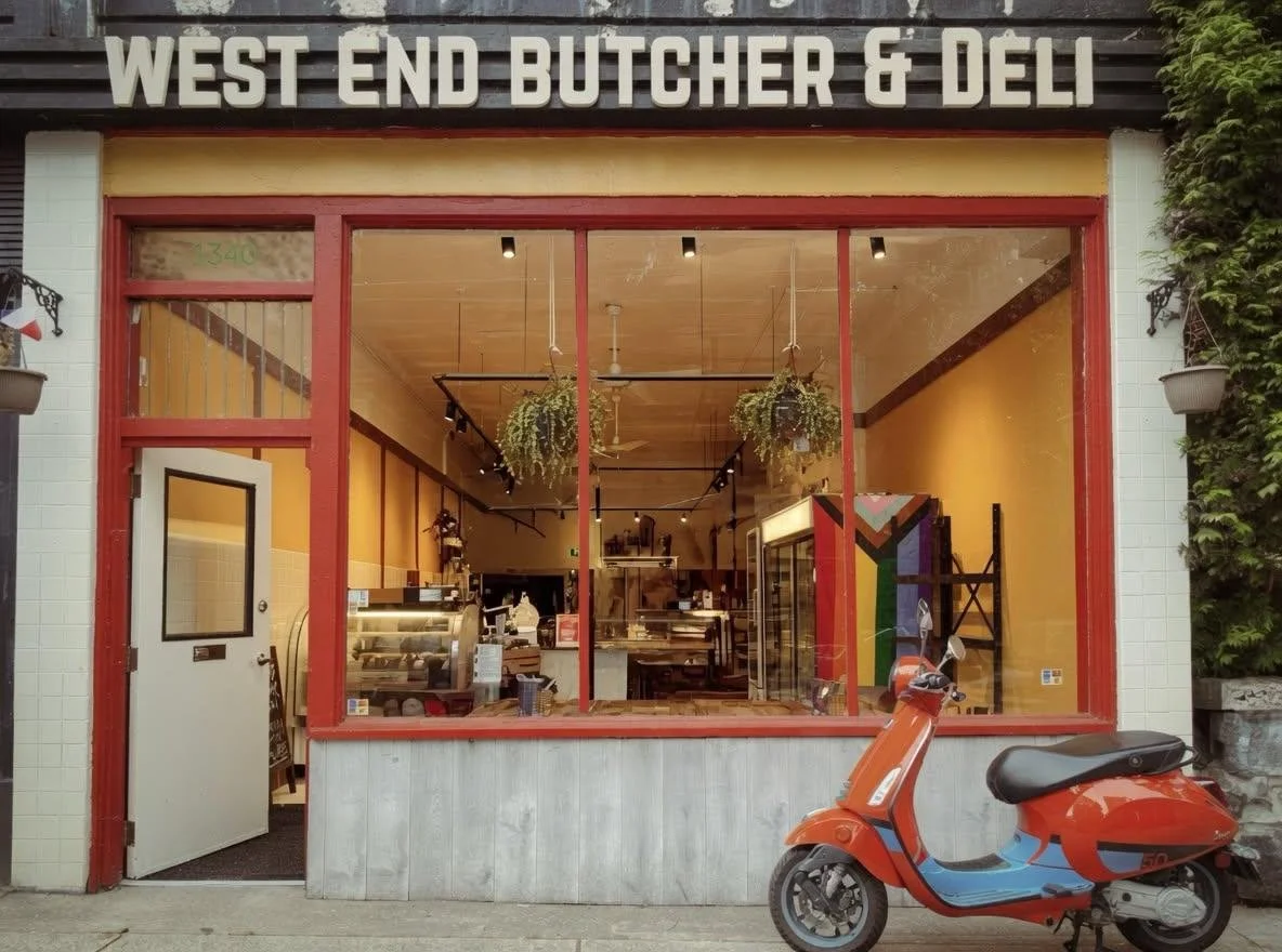 storefront of West End Butcher & Deli with a large front window, an orange scooter parked outside, and hanging plants inside visible through the window.