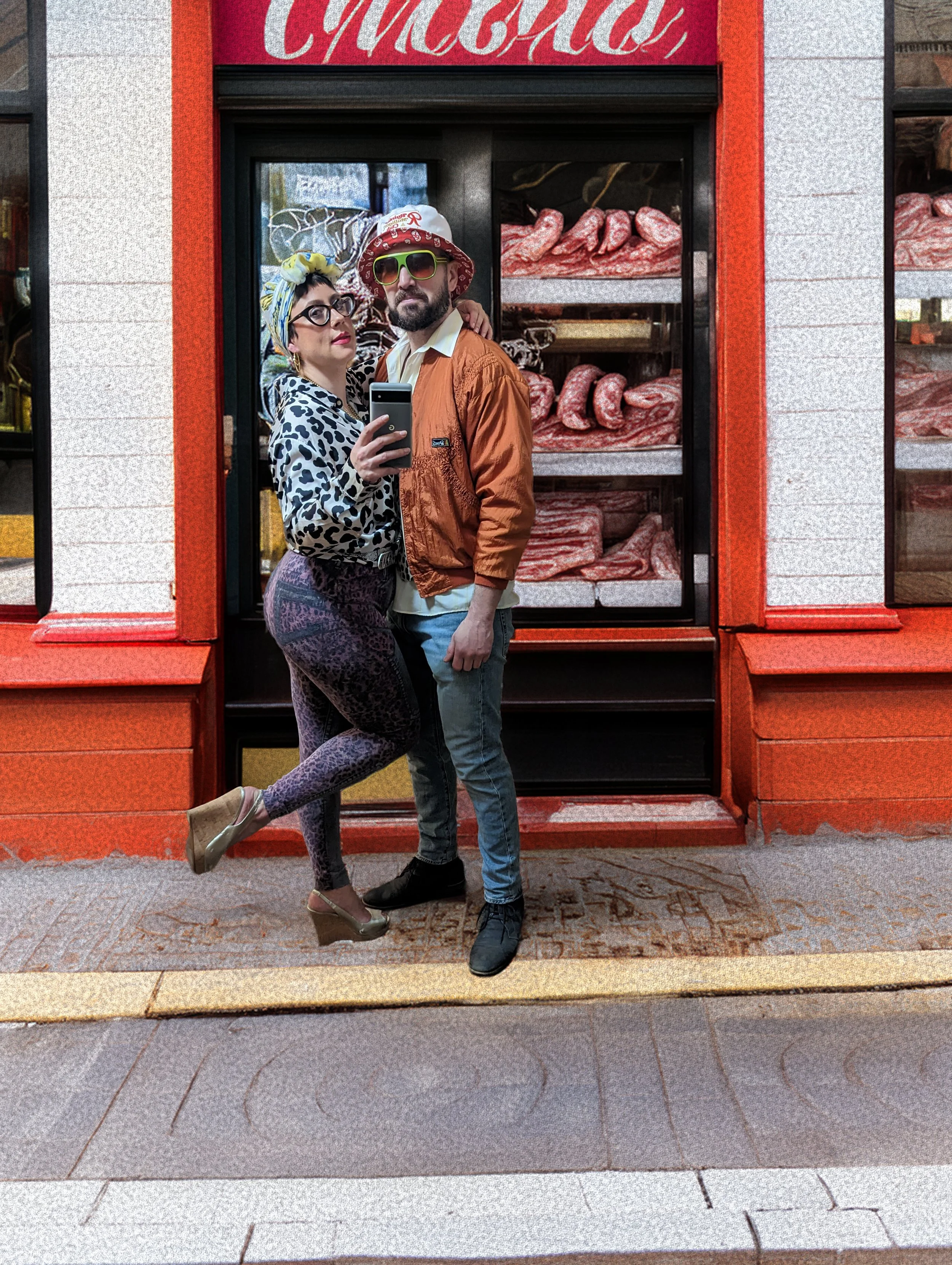 A man and woman taking a selfie in front of a butcher shop window with displayed meats, dressed in colorful, vintage-inspired clothing and accessories.