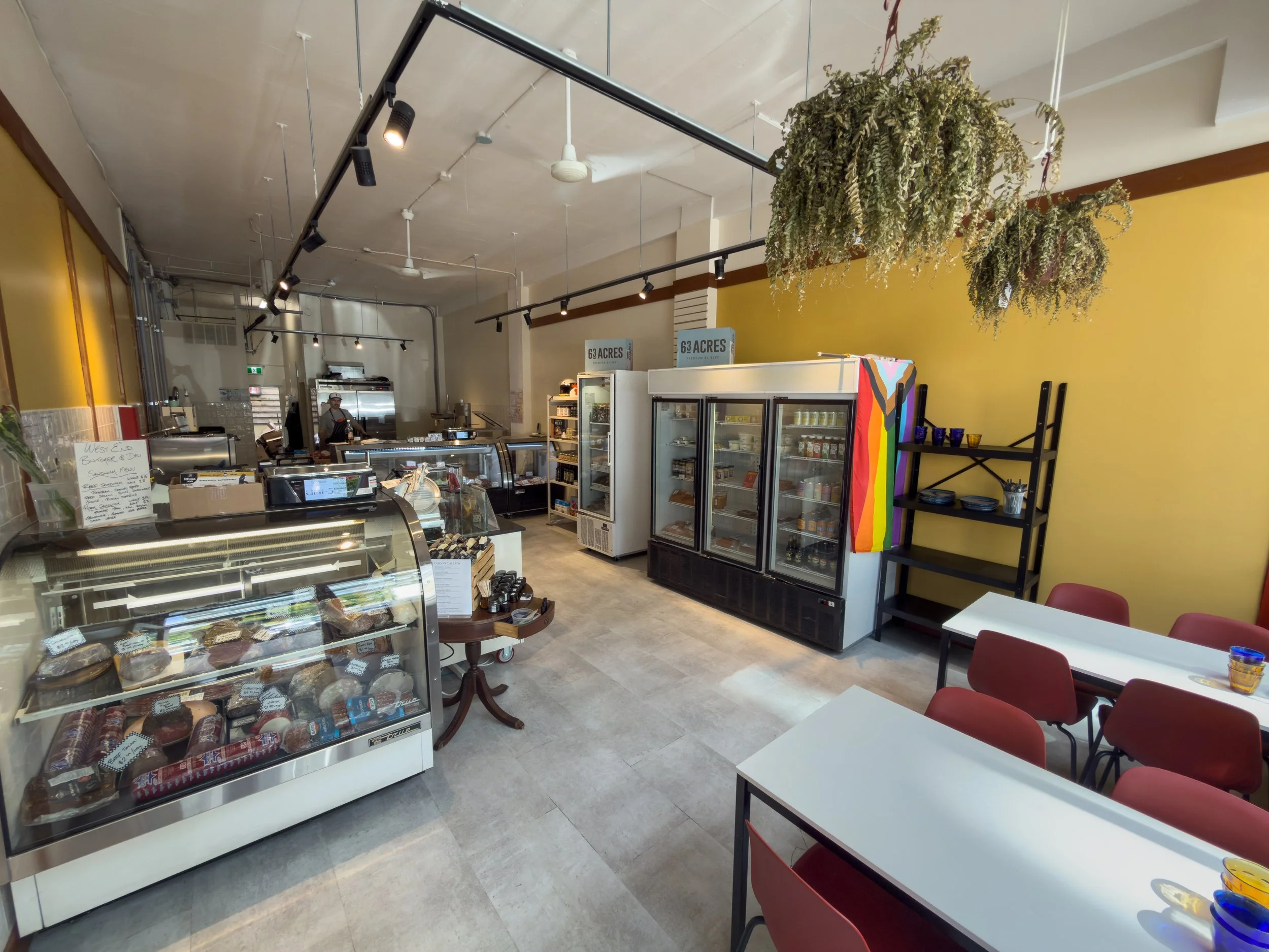 Interior of a bakery or cafe with shelves of baked goods, a counter, and tables with red chairs. Hanging plants and lighting fixtures are visible on the ceiling.