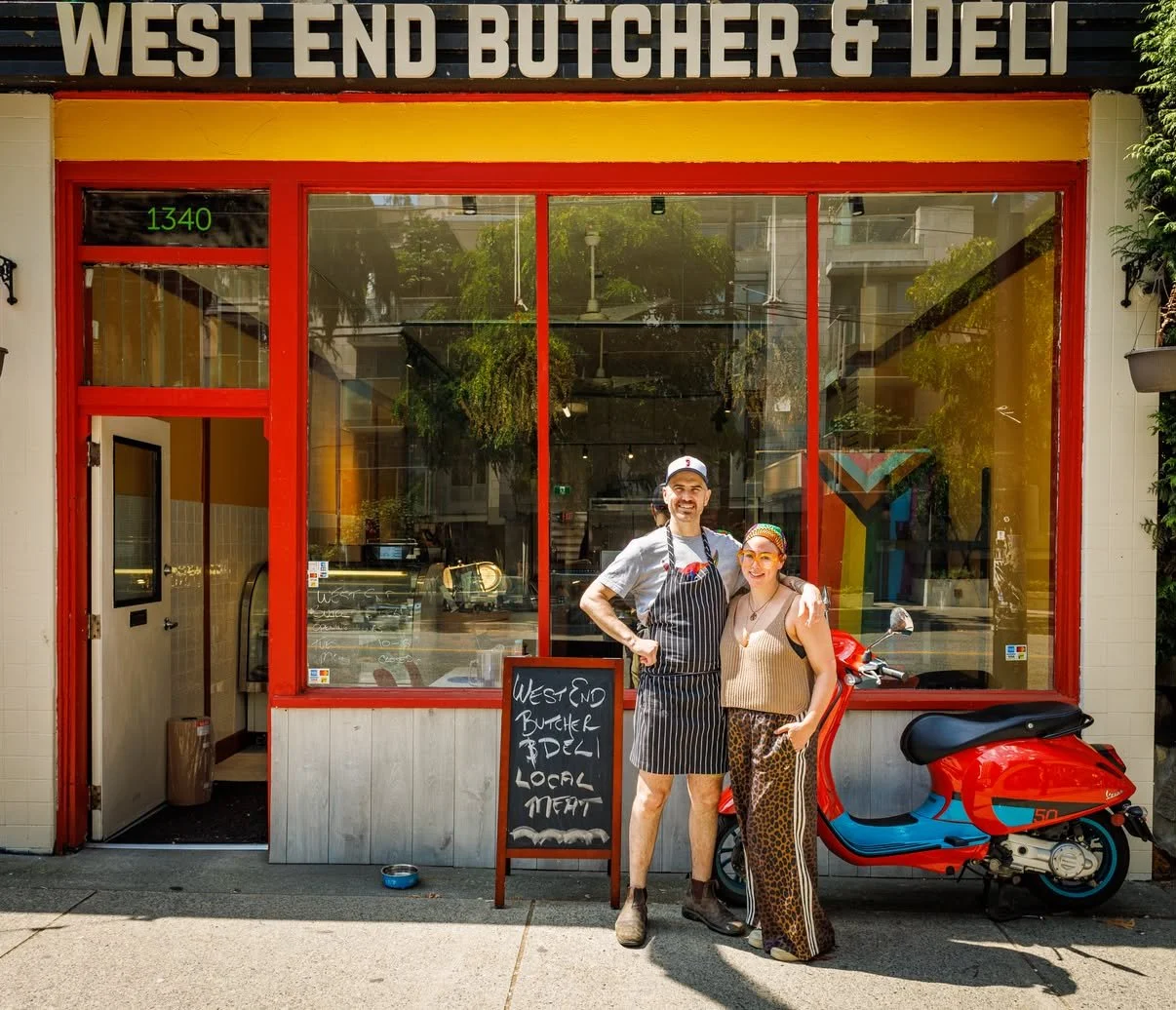 A man and woman stand outside a butcher shop called 'West End Butcher & Deli' with a red and yellow exterior, a chalkboard sign, and a red scooter parked nearby. They are smiling and posing for the photo.