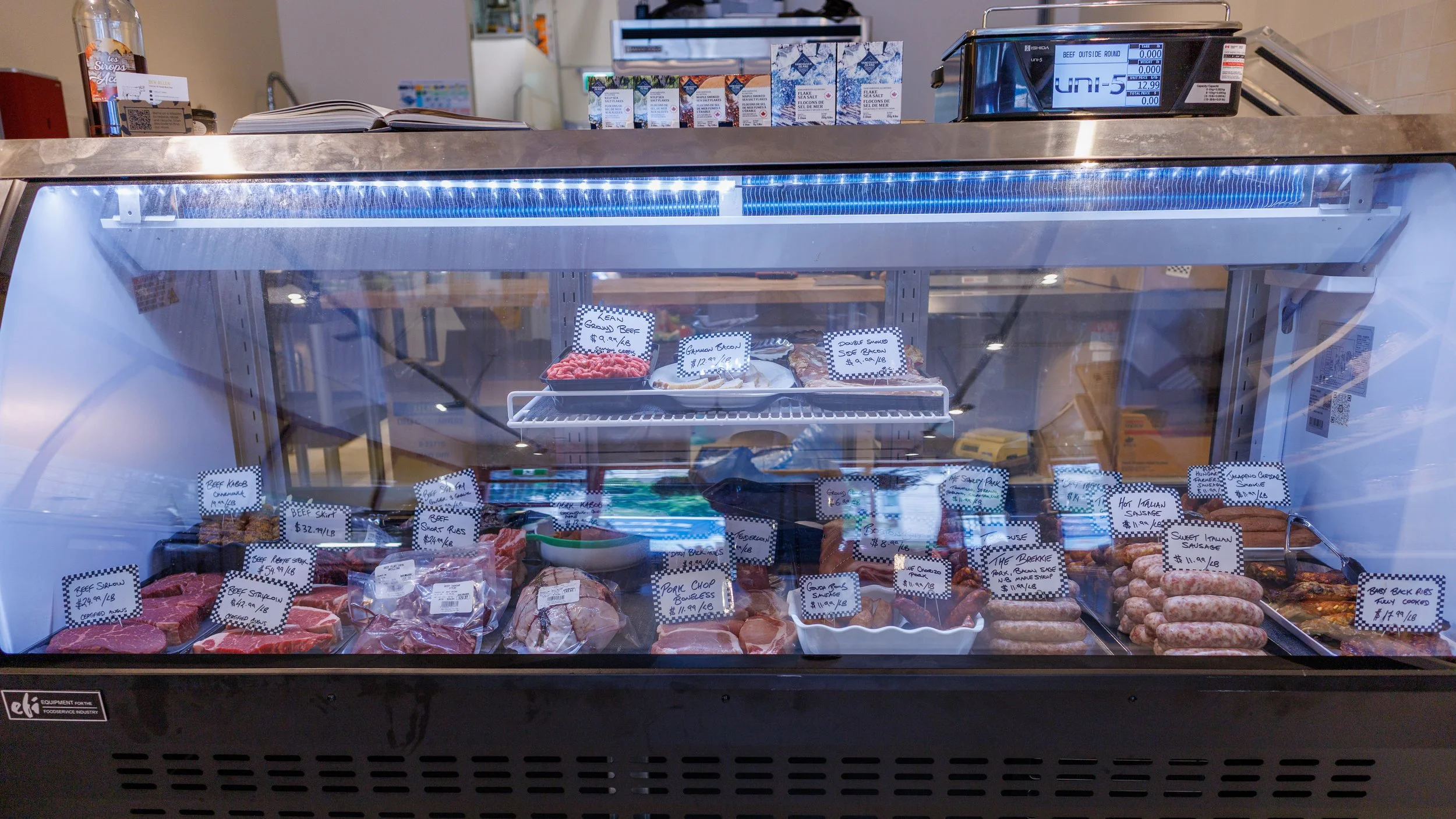 A glass display case filled with various cuts of meat, including beef and sausage, with handwritten labels. The top of the case shows a counter with smaller packaged products, a cookbook, and a digital scale.