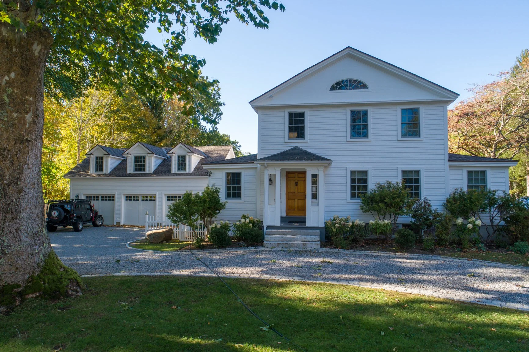 A large white two-story house with a wooden front door, set amidst green trees and bushes. There is a gravel driveway with a black vehicle parked to the side, and a small white picket fence near the bushes. The sky is clear and blue.