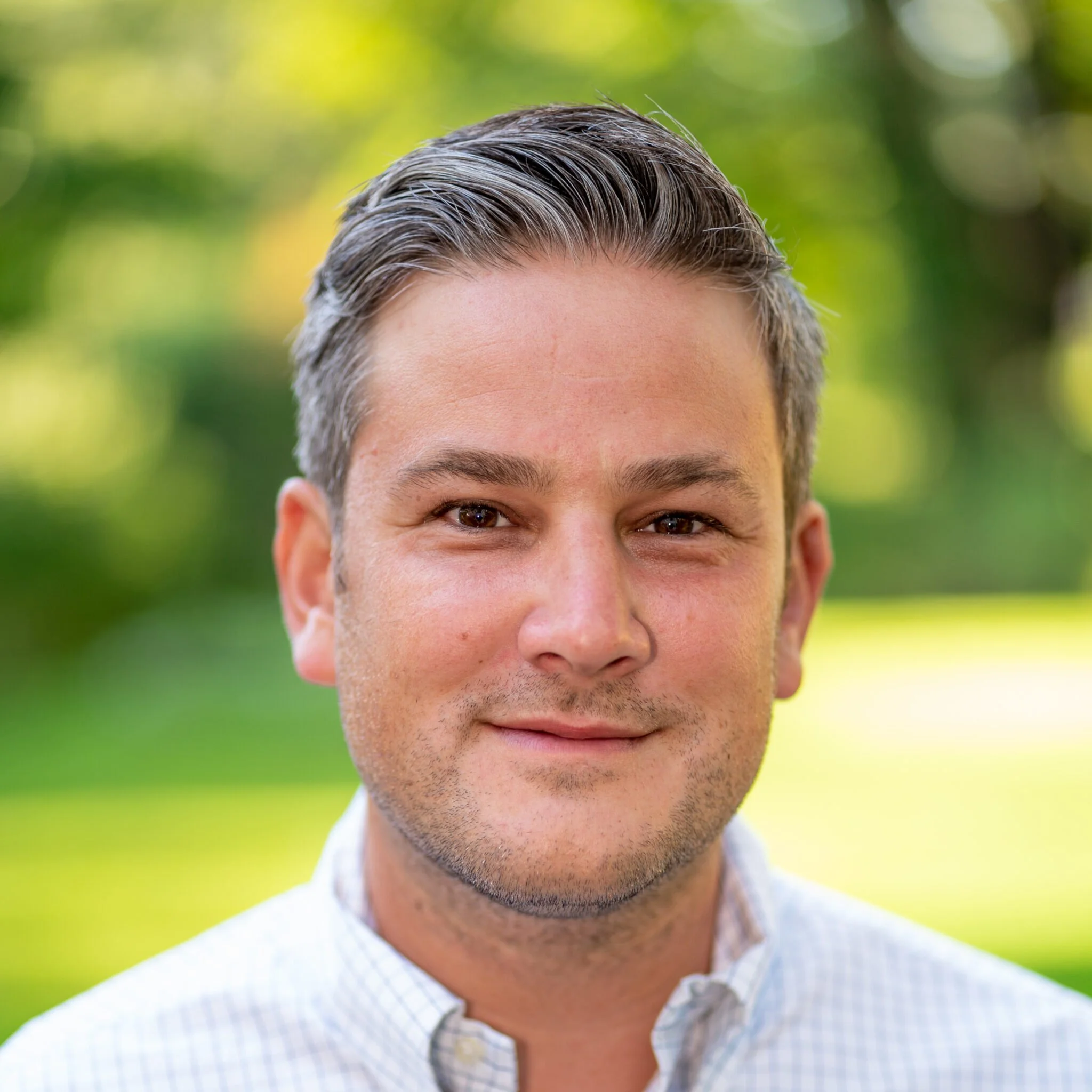 A close-up portrait of a man with short dark hair, smiling, outdoors with blurred green foliage in the background.