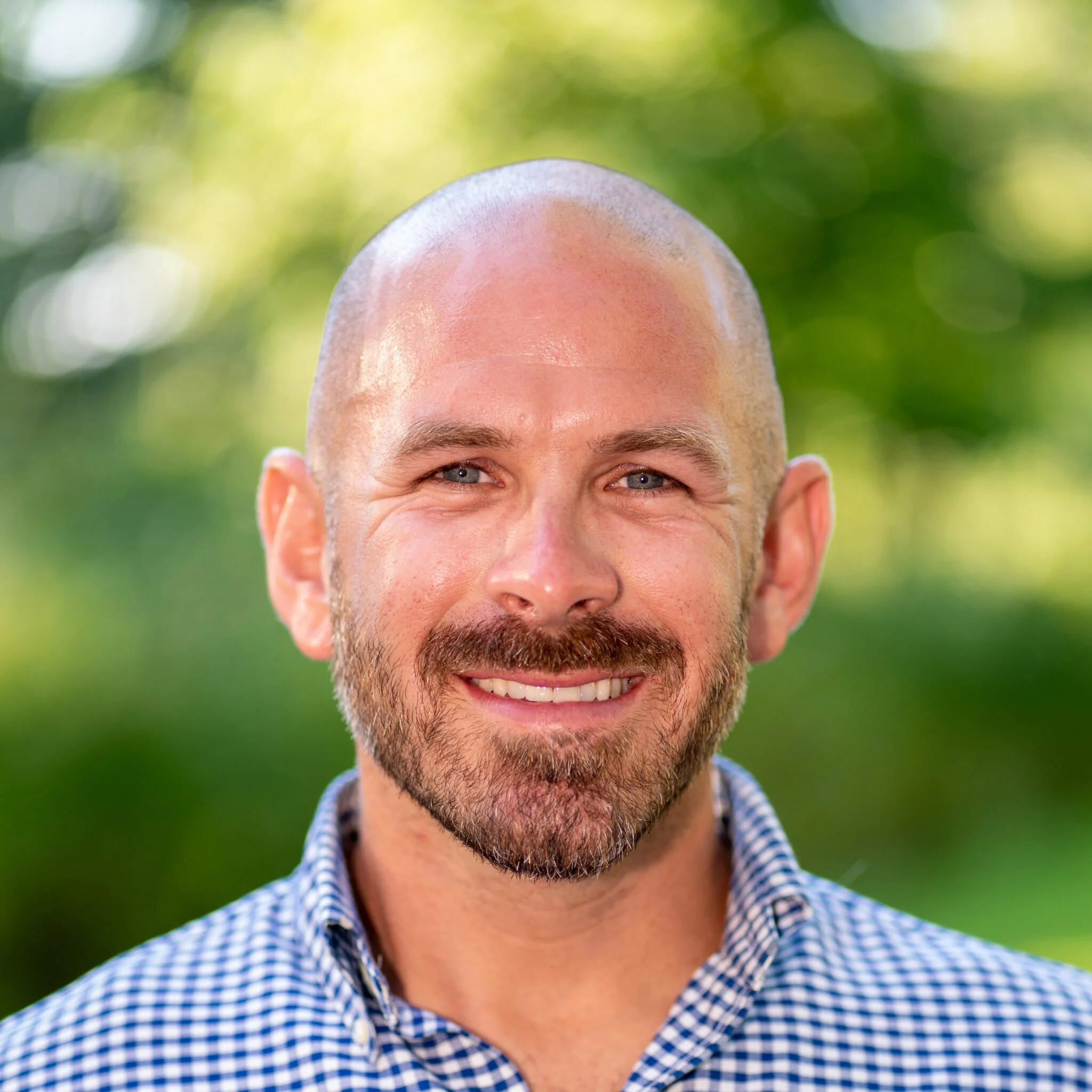 Close-up of a smiling man with a shaved head and beard, wearing a blue checkered shirt, outdoors with green blurred foliage background.