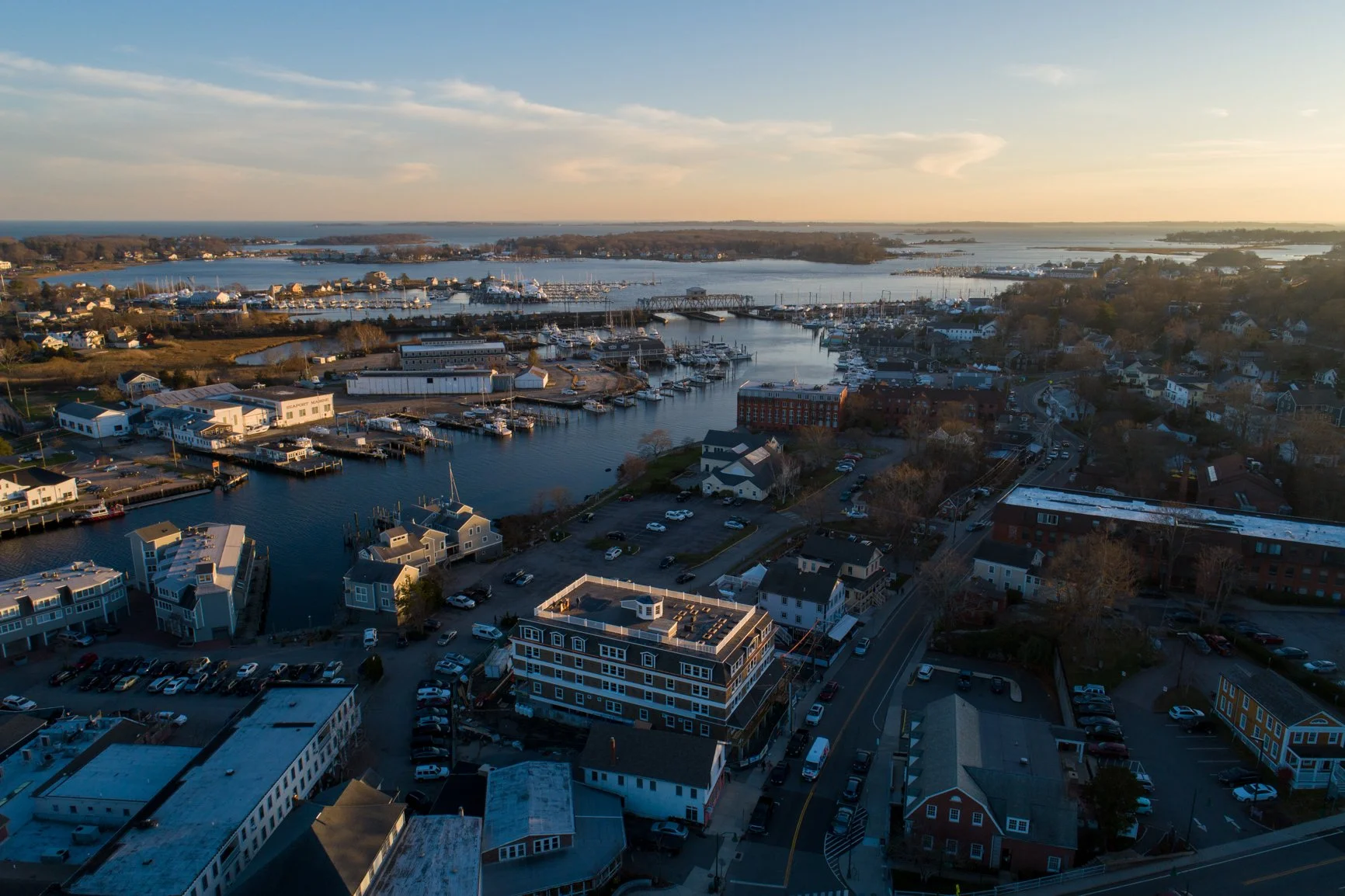 An aerial view of a harbor town at sunset, showing boats docked in the marina, residential houses, and a mix of commercial buildings along the waterfront with calm water and a partly cloudy sky.