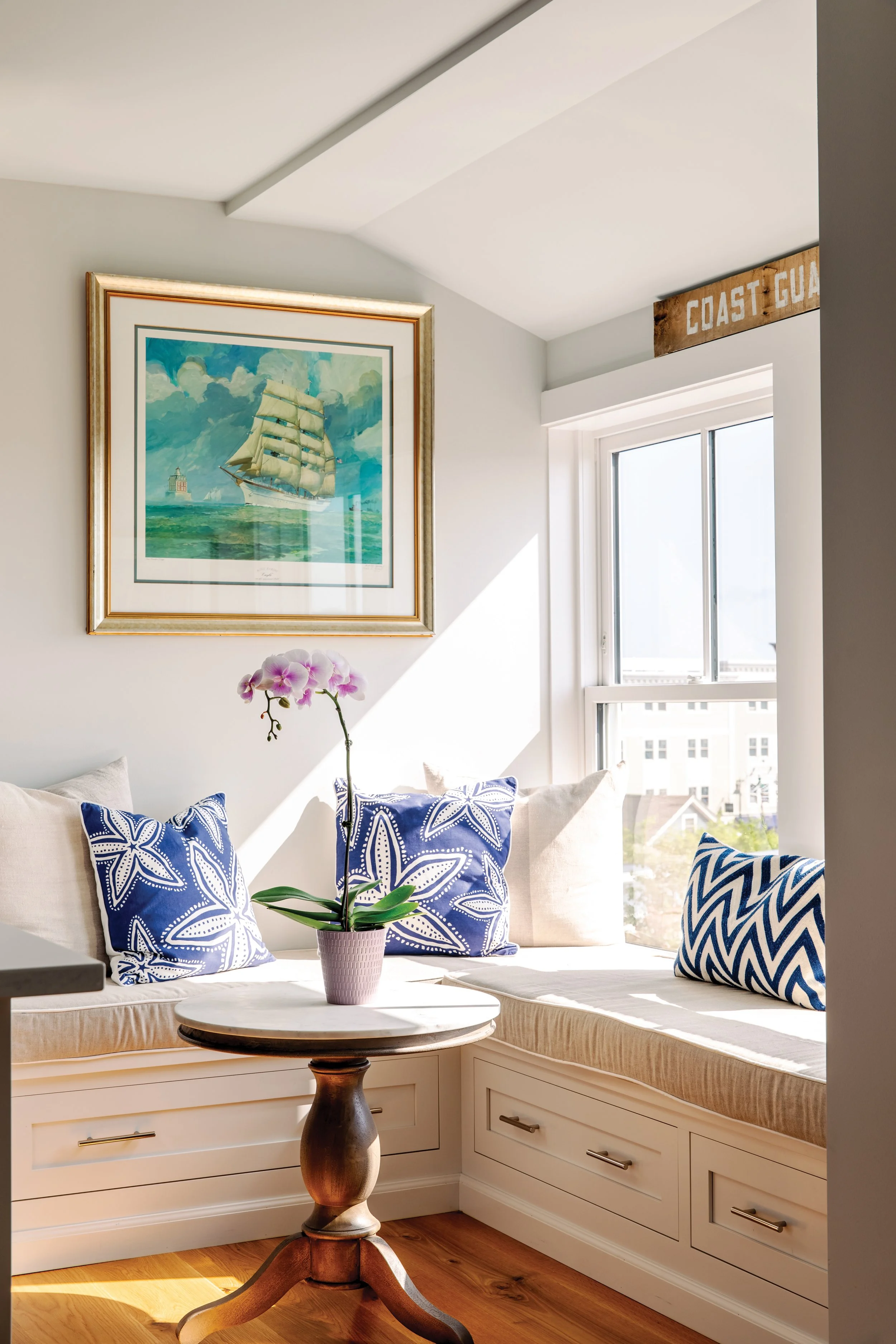 Sunlit coastal-themed window nook with built-in white bench, blue patterned pillows, a white cushion, a small round table with a pink orchid, artwork of a sailing ship on the wall, and a 'Coast Guard' sign above the window.