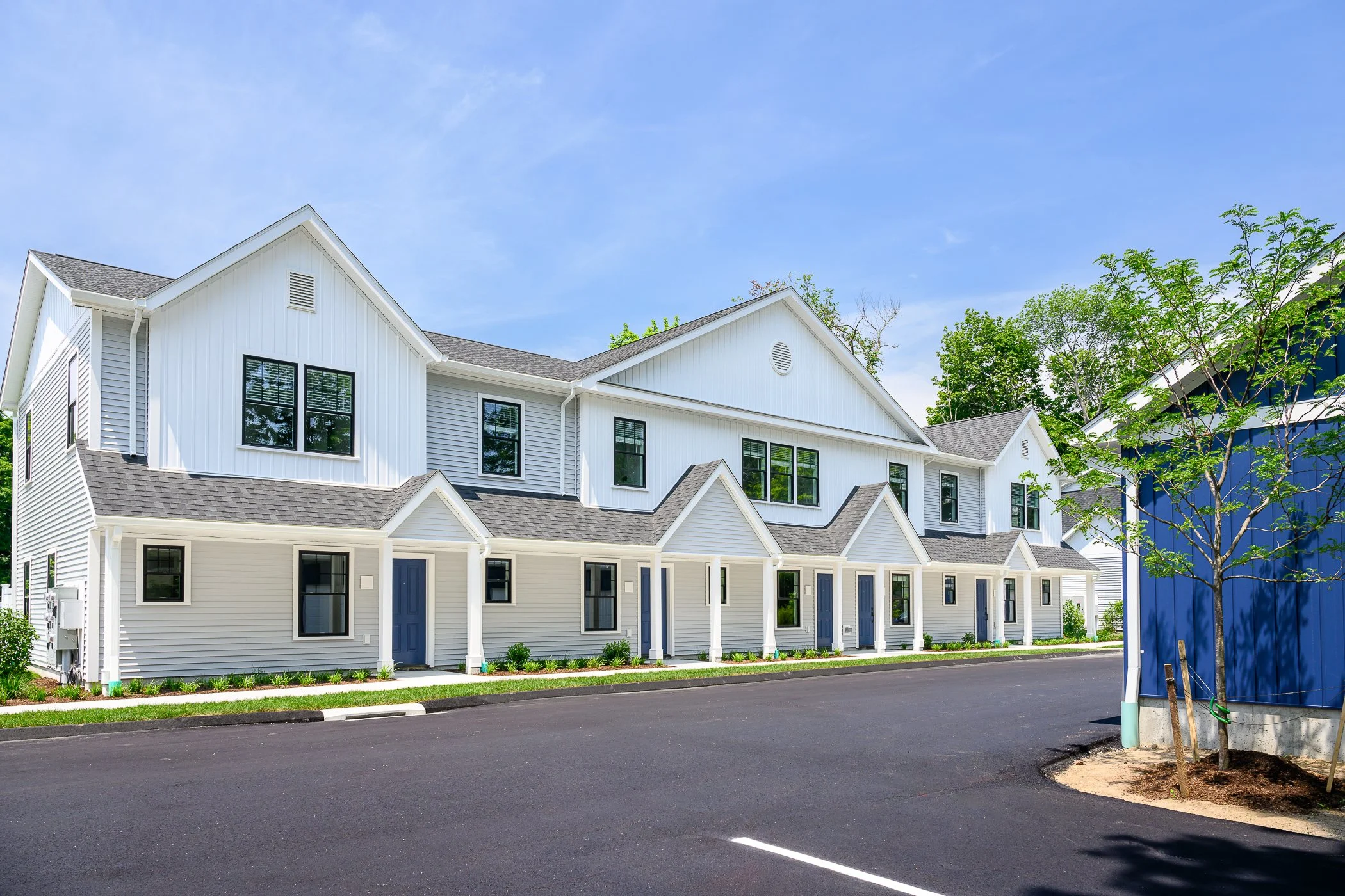 A modern white multi-family residential building with multiple entrances, situated along a paved street with a single young tree, under a clear blue sky.