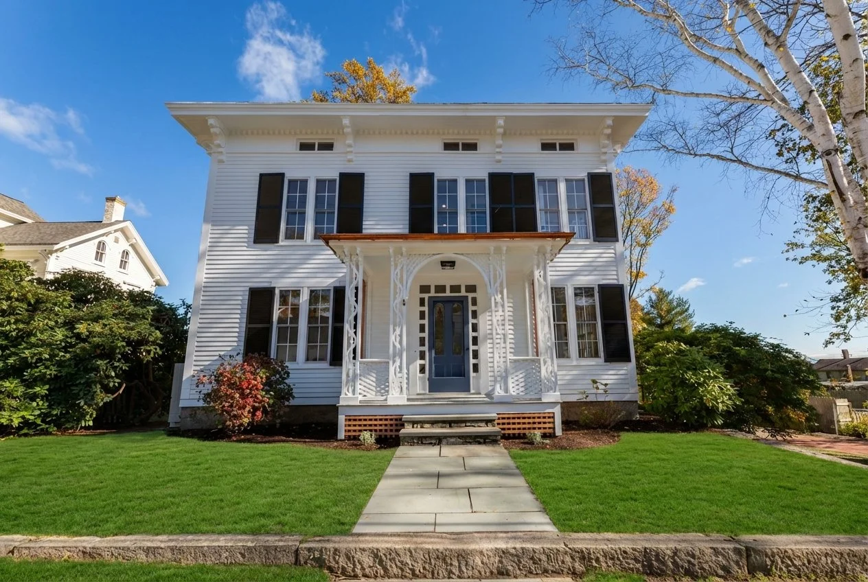 A two-story white house with black shutters, a front porch with decorative columns, and a small set of stairs leading to the front door. The house has a well-maintained lawn and garden, with a tree on the right side.