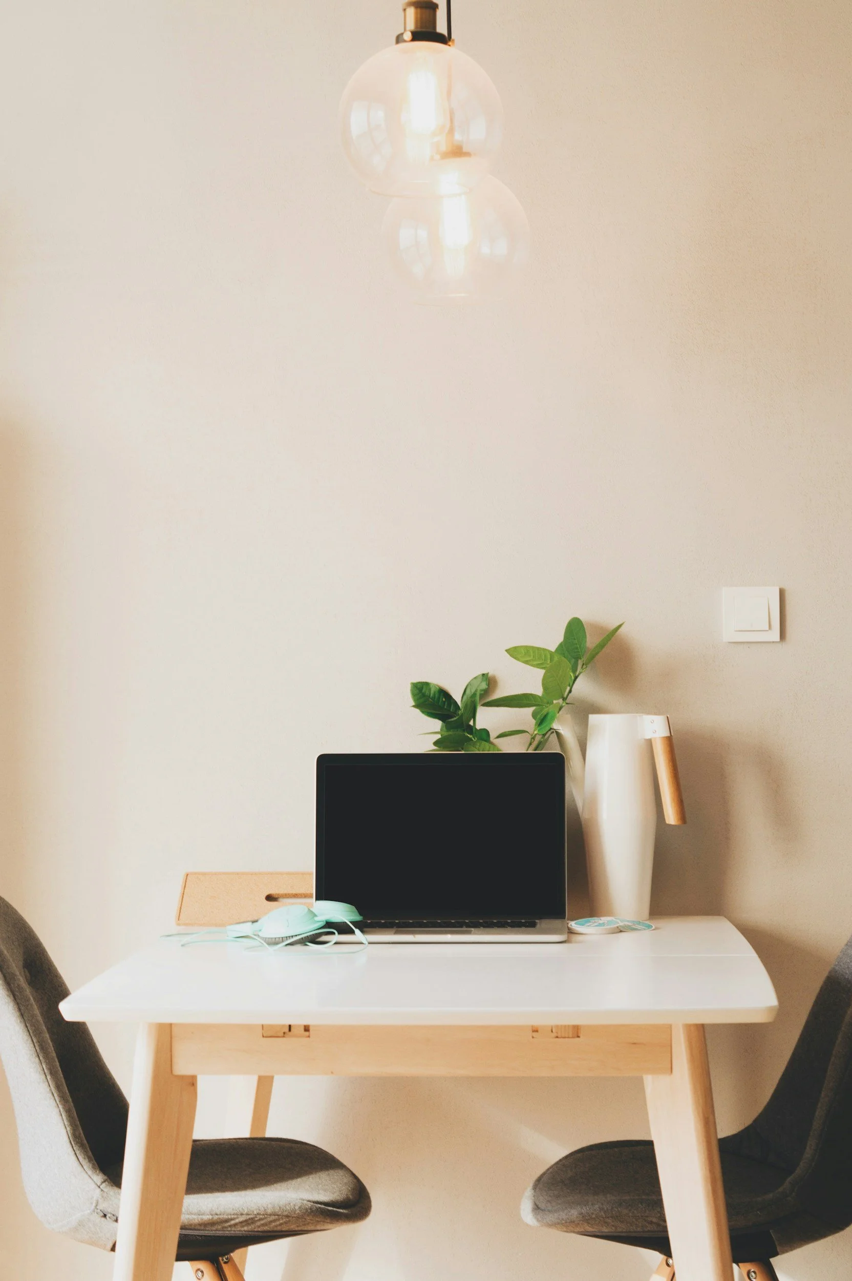 A modern home workspace with a white table, two black chairs, a closed laptop, a face mask, a green plant, a white pitcher, and a decorative item on a wall with a white switch.