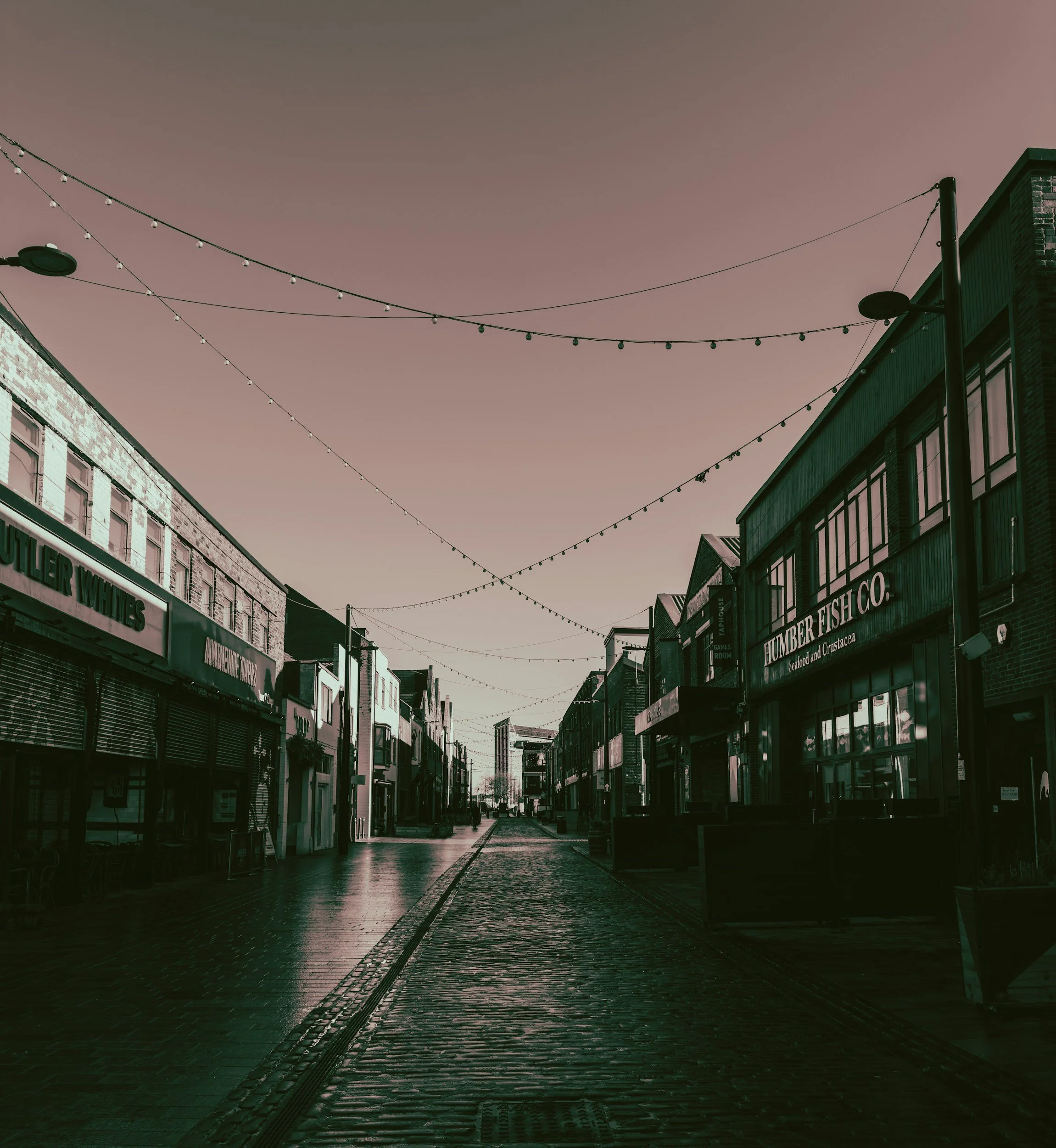 Empty street with shops and string lights overhead, captured during sunset or dawn.