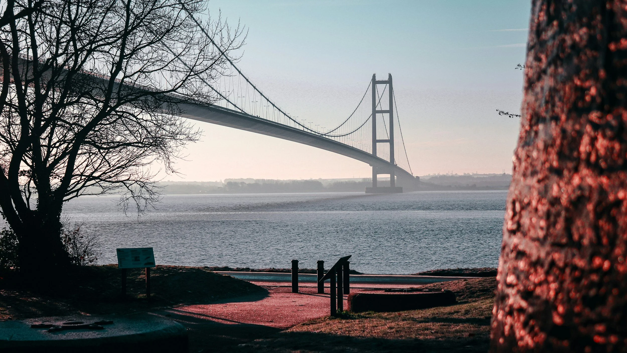 A view of a large suspension bridge over a body of water, with trees in the foreground and a pathway along the water's edge.