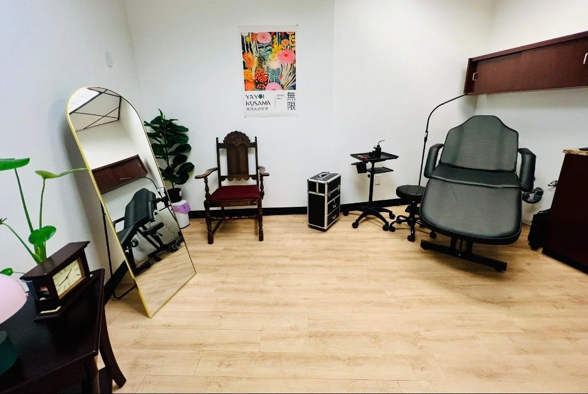 A tattoo studio with a desk chair, a framed mirror leaning against the wall, a potted plant, a vintage chair, a small table with items, a poster on the wall, and various furniture and decor.