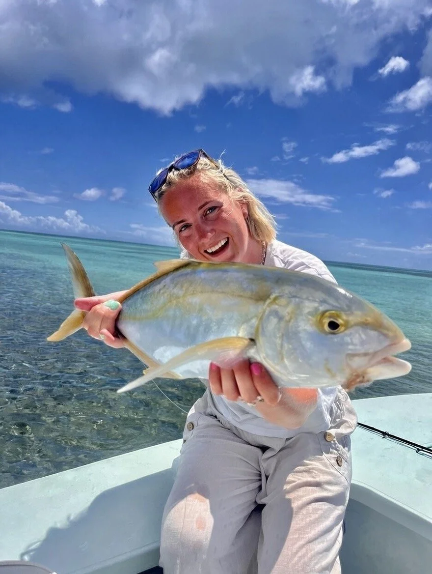 A woman in a white shirt and beige pants holding a large fish on a boat in a clear blue ocean with partly cloudy sky.