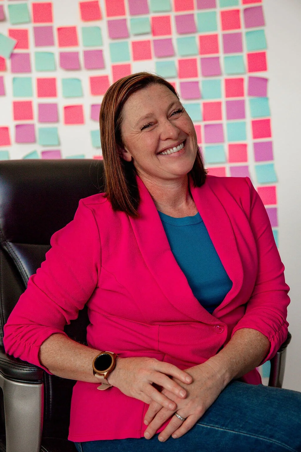 Liz Chapman, podcast producer and strategist, is sitting in her chair smiling at the camera. She is wearing a turquoise shirt with a bright pink blazer, and on the wall behind her are a mirage of pink, purple and blue post it notes