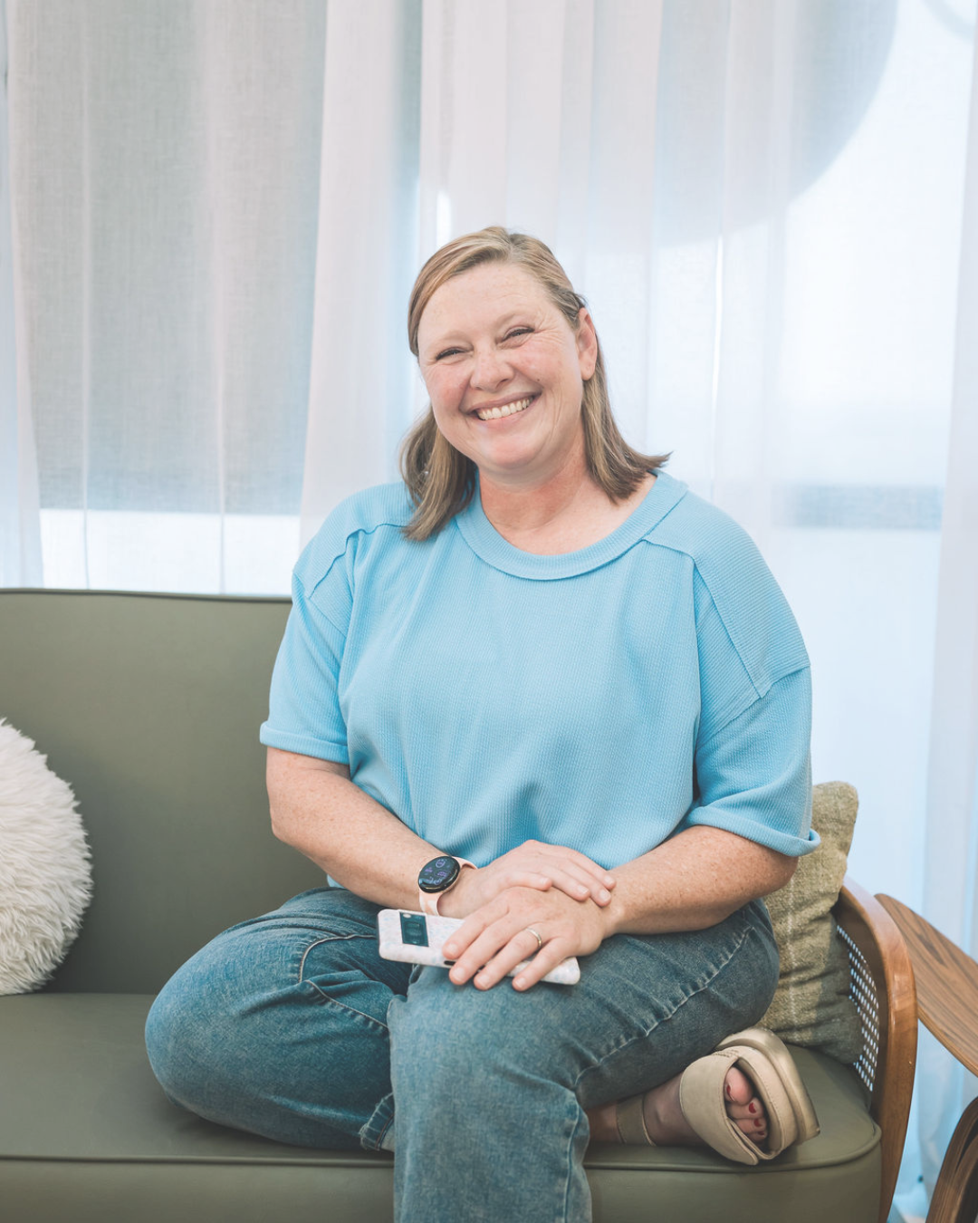 Liz Chapman smiling brightly at the camera in a blue shirt with her hands on her lap and you can see her phone under her hands