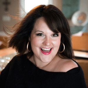 Andee Hart, host of the She Sells Differently Podcast, smiling at the camera with a black sweater on and fabulous gold hoop earrings