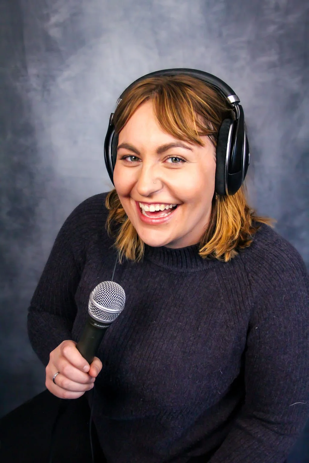 Jess Schmidt, podcast editor, smiling at the camera with headphones on and holding a microphone. 