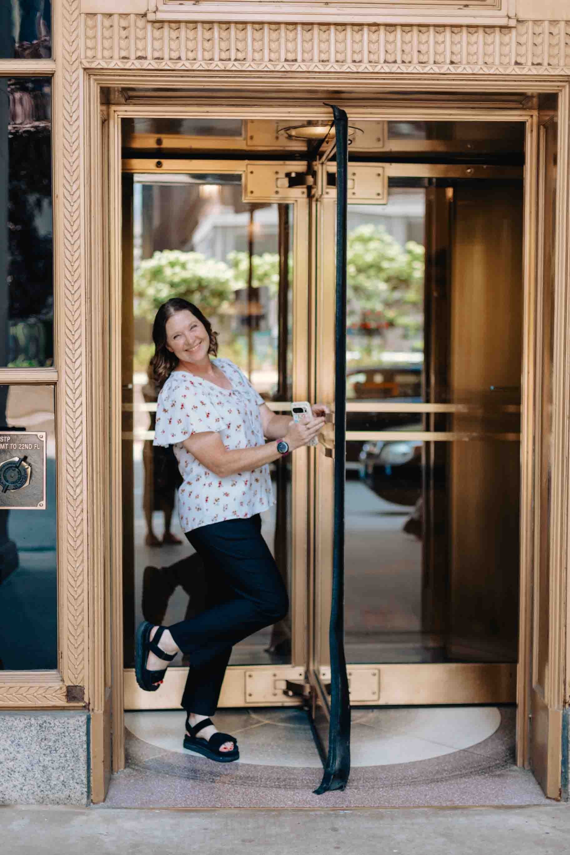 Liz Chapman is a podcast coach and she is walking out of a revolving door smiling at the camera with her foot in the air in downtown Chicago