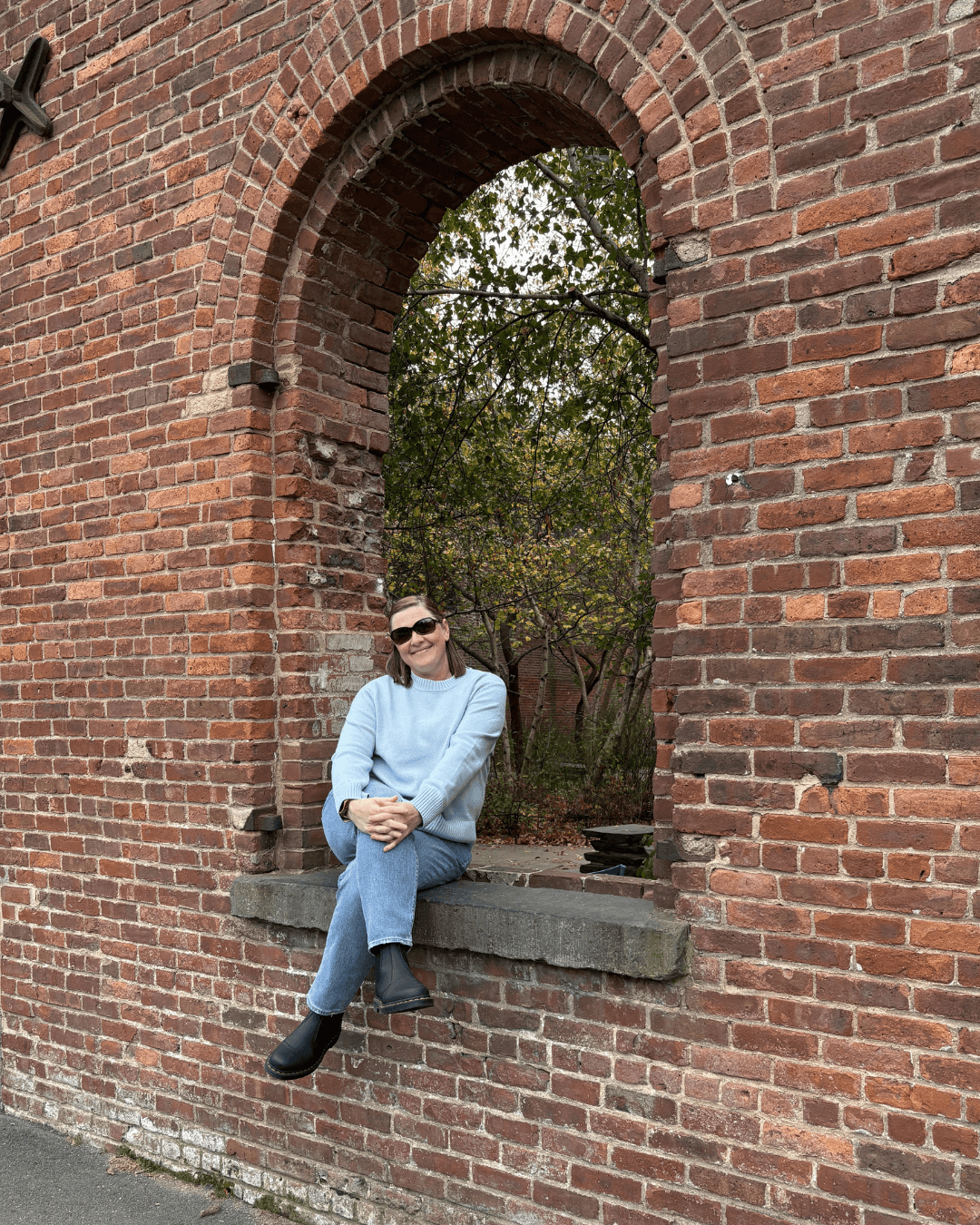 Liz Chapman is a podcast coach and she is sitting on the windowsill in Brooklyn, NY on a fall day and smiling at the camera.