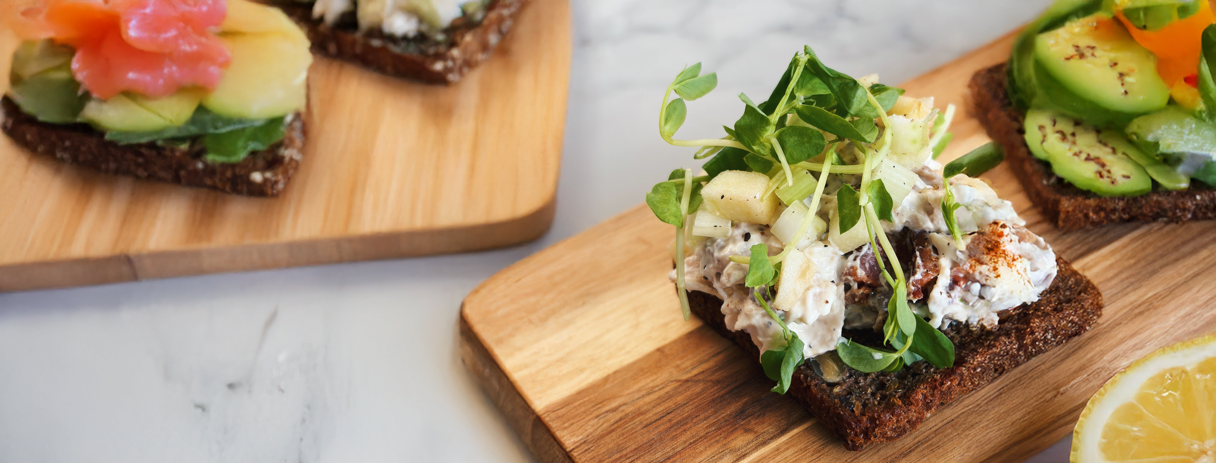 Close-up of three open-faced sandwiches on wooden boards, topped with various vegetables and spreads, with a lemon wedge in the corner.
