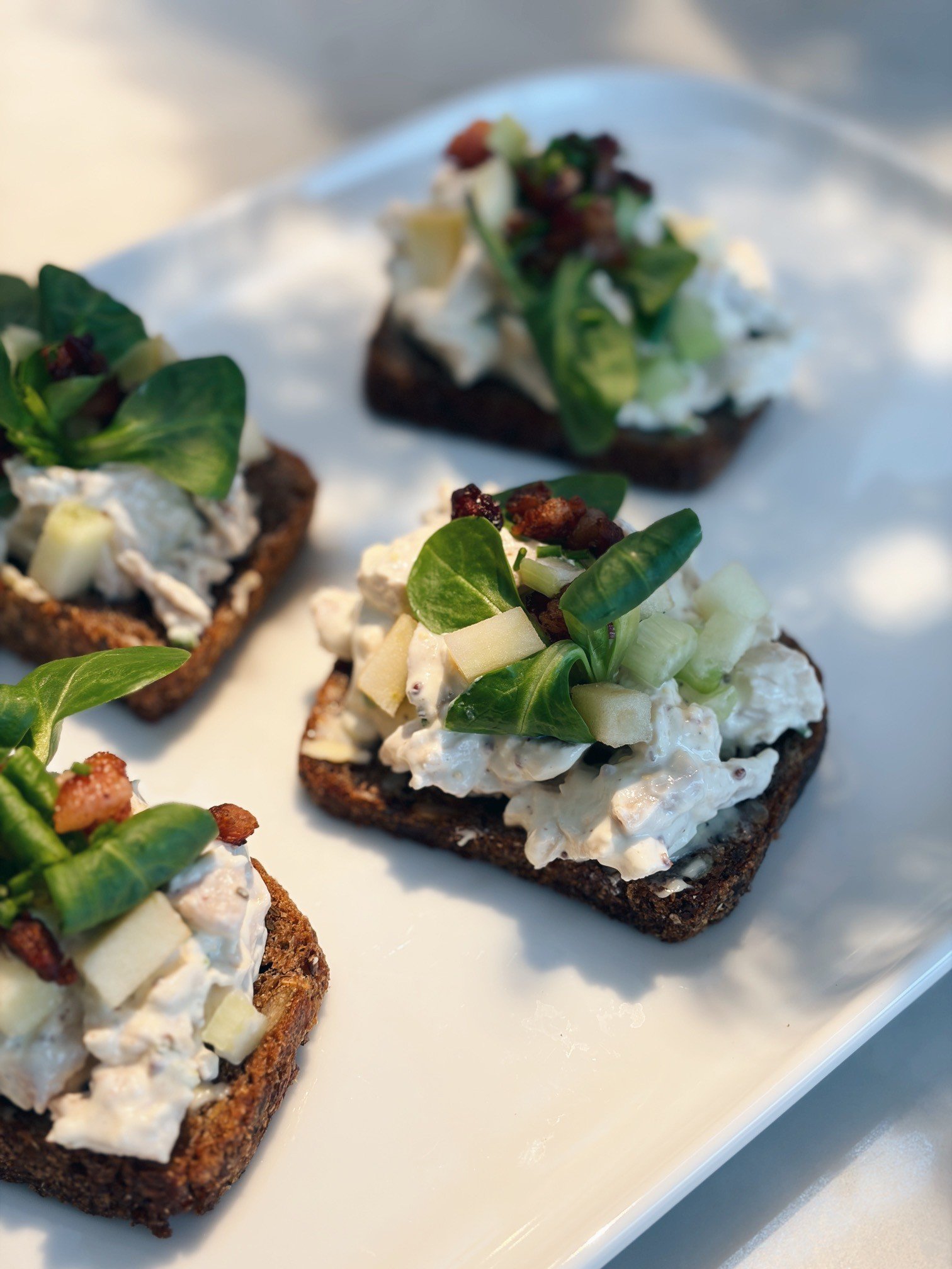Three pieces of open-faced sandwich with white spread, green leafy garnish, and bacon bits on dark toasted bread, served on a white platter.